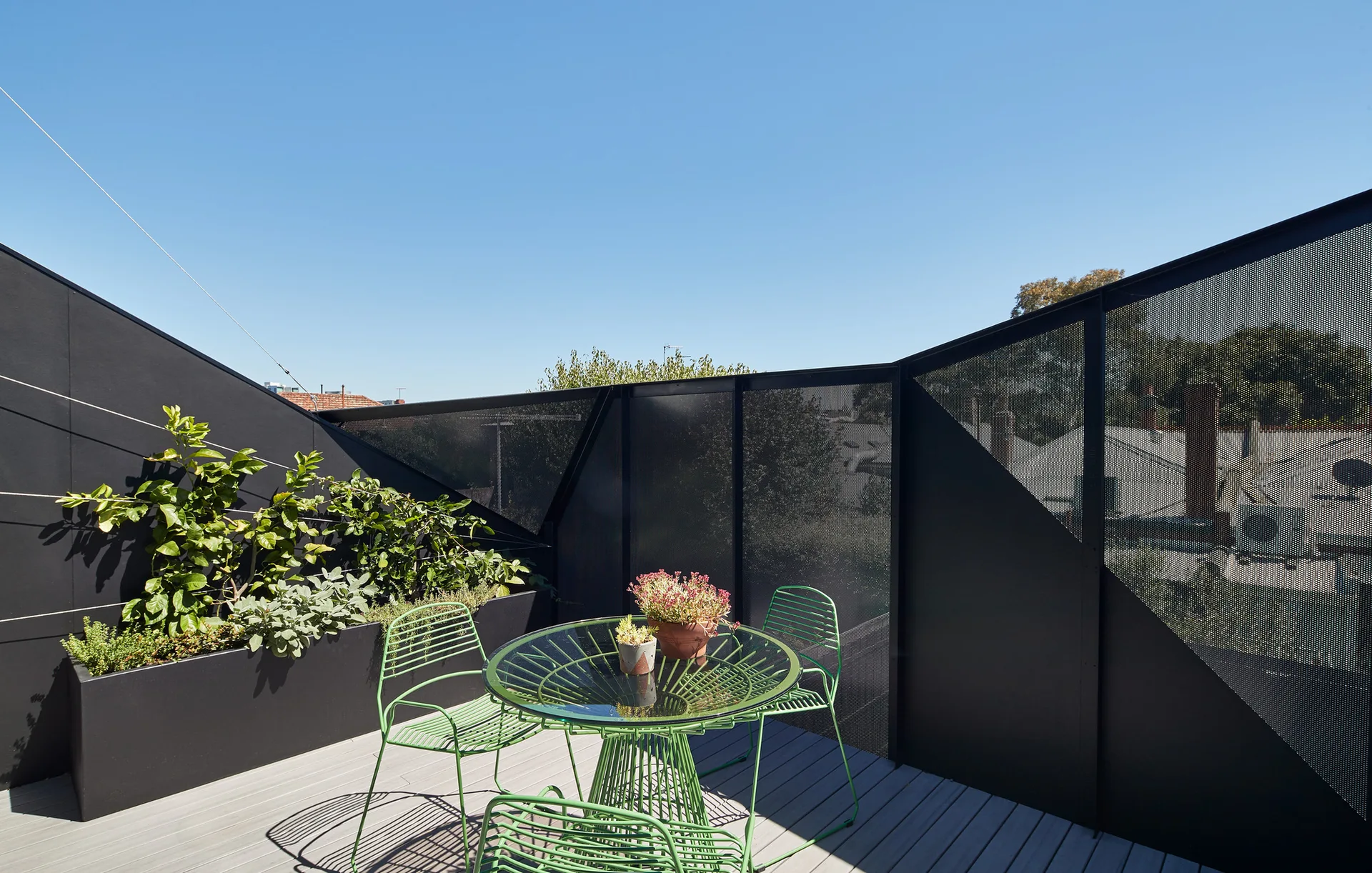 A grey plank rooftop deck features a green wire metal bistro set and a black perforated metal privacy screen next to a planter box.