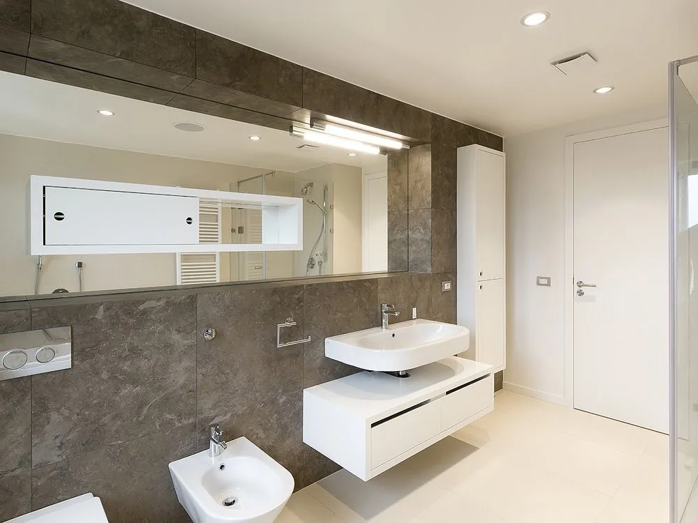 A modern bathroom features a dark grey stone tiled wall, a white floating bidet, and a wall-mounted vanity under a large mirror.