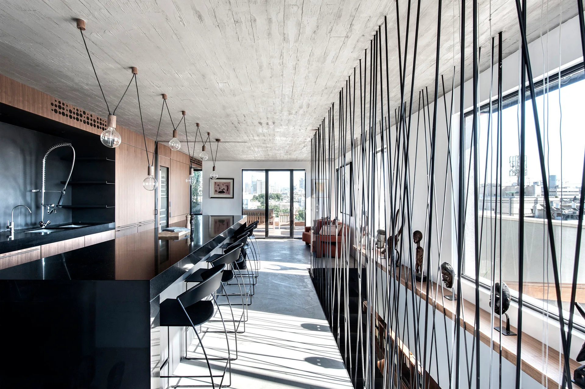 A modern kitchen with a long black island, wood wall cabinetry, a textured concrete ceiling, and a vertical metal rod room divider.