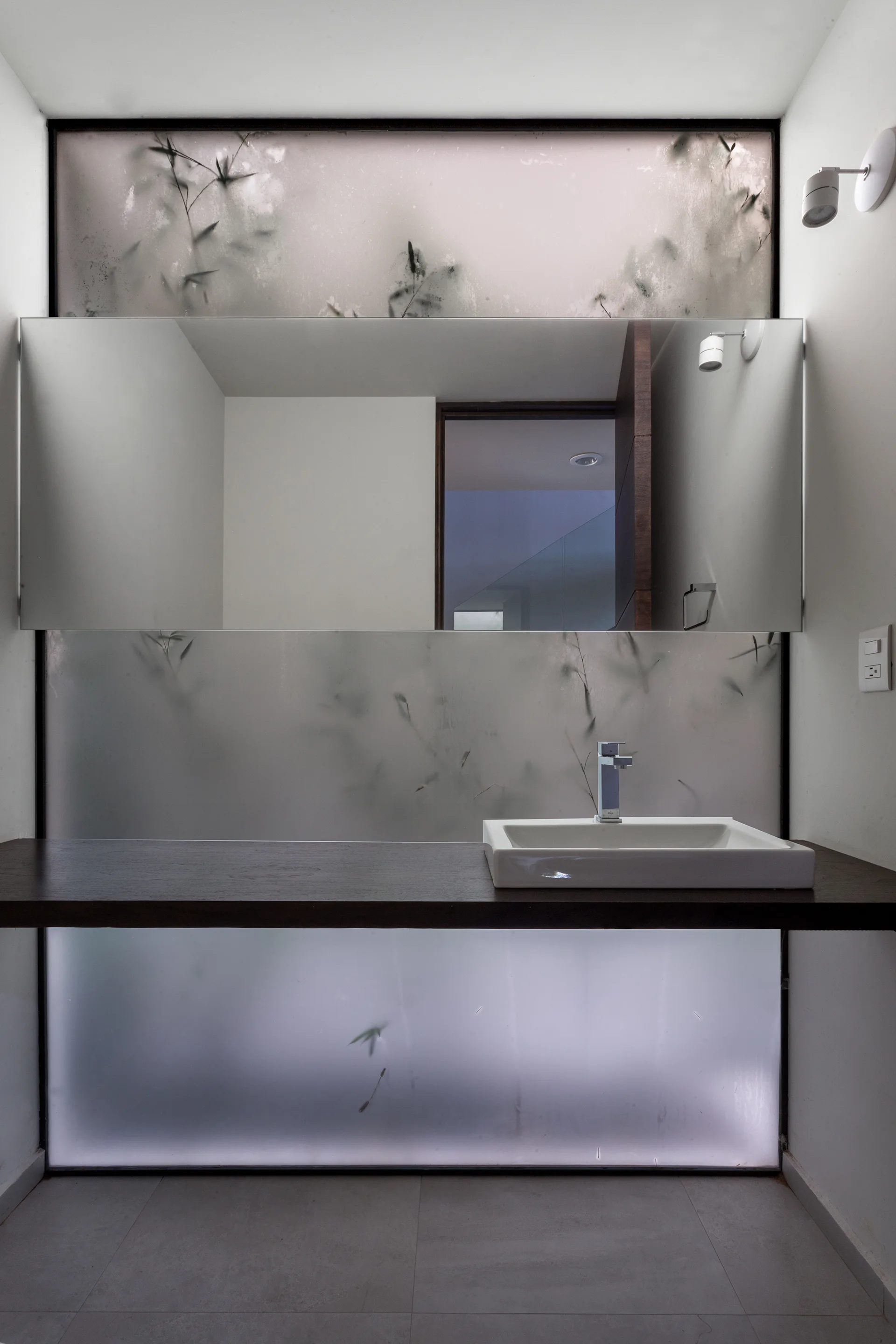 A minimalist bathroom vanity features a floating dark wood shelf, white sink, and large mirror set against backlit frosted glass panels with plant silhouettes.