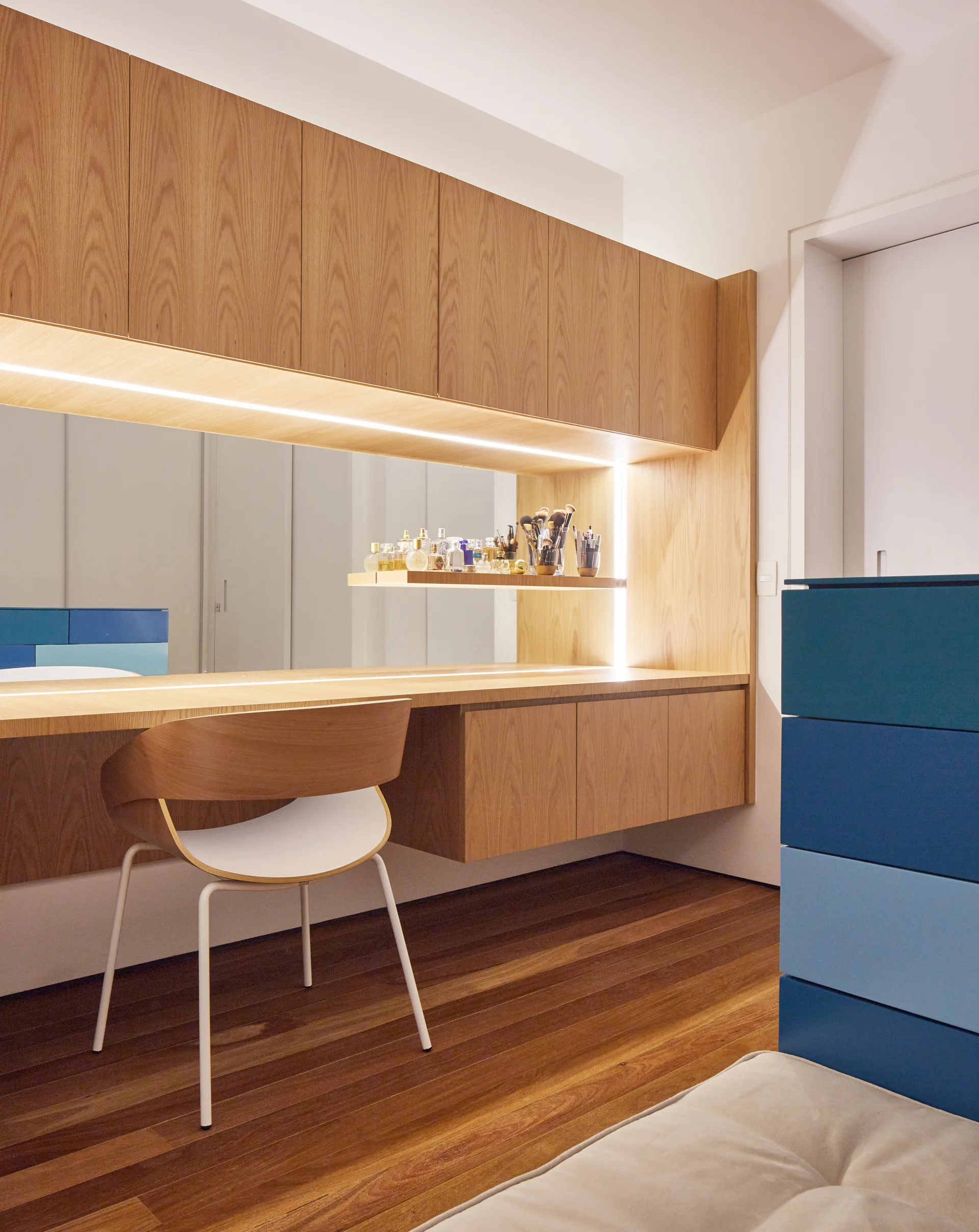 A modern dressing room features a built-in wooden vanity with a large mirror, integrated LED strip lights, and a curved wooden chair.