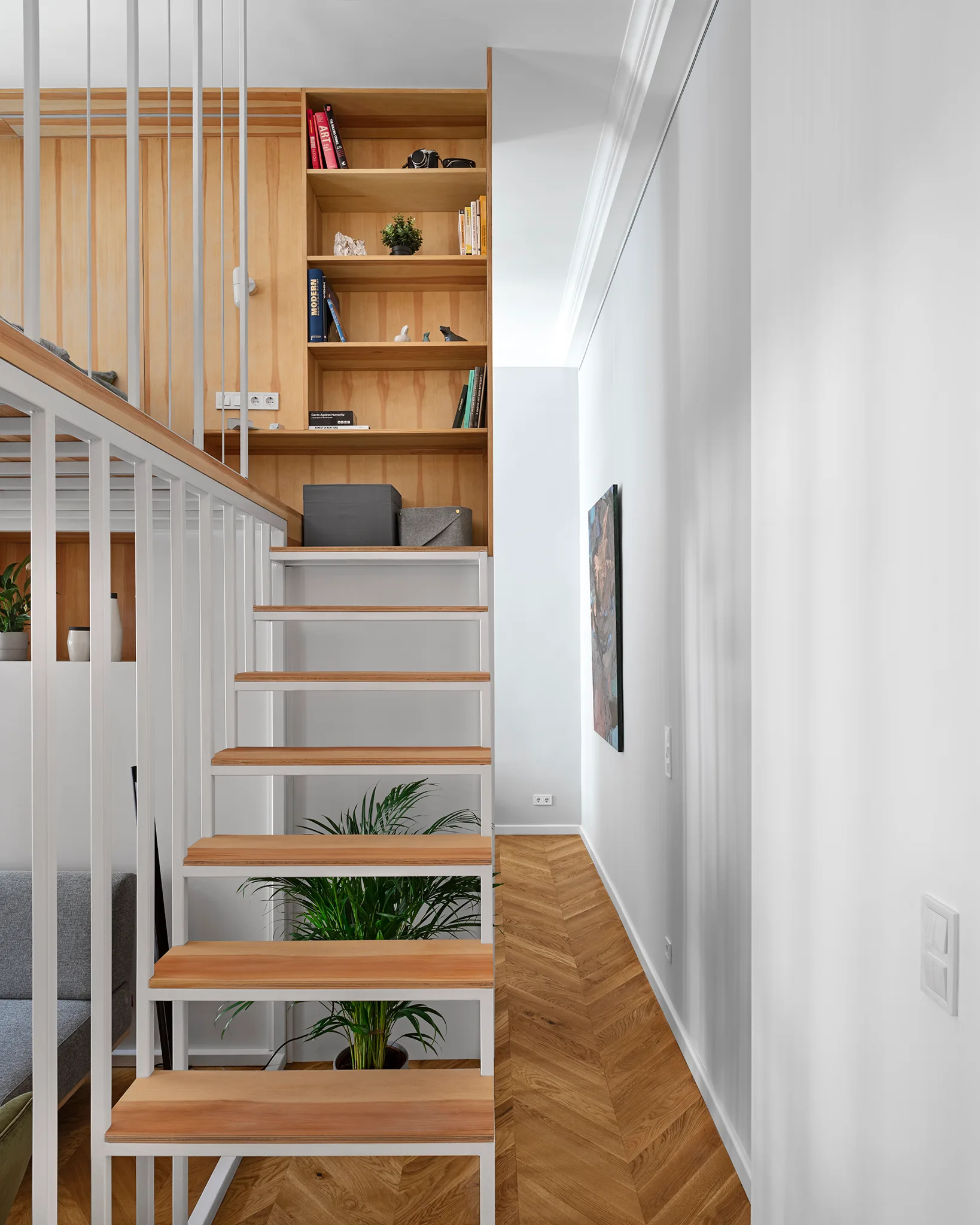 A modern floating staircase with light wood treads and white railings stands next to built-in wooden shelving over herringbone hardwood floors.