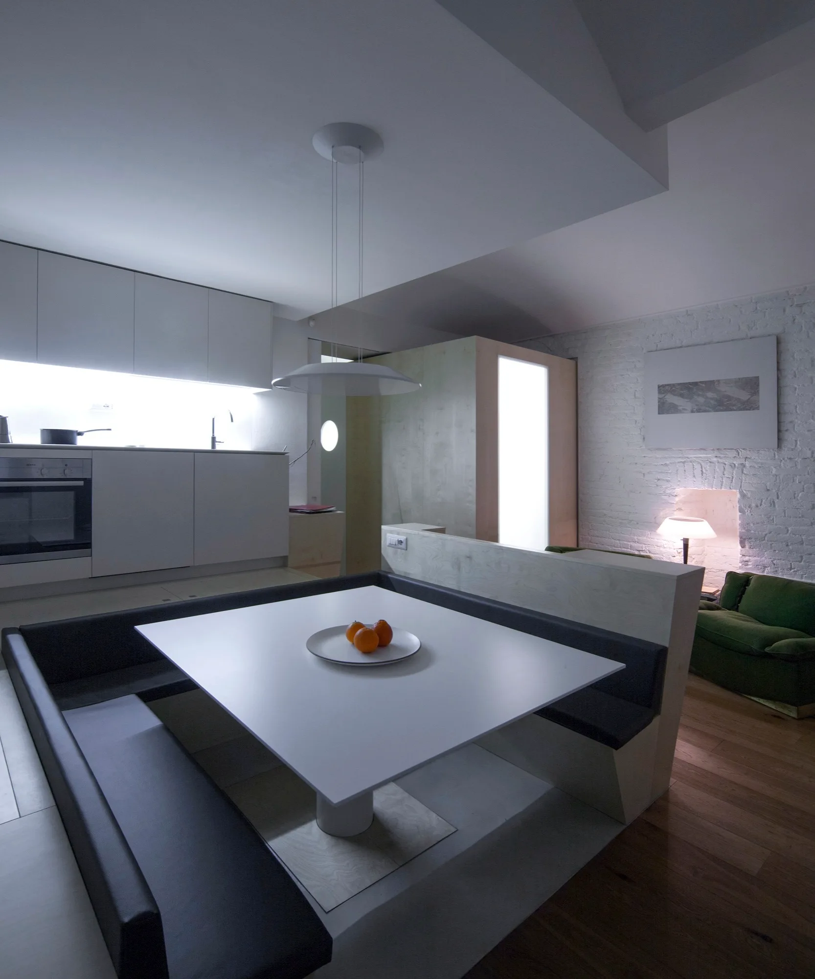 A modern kitchen space features a built-in U-shaped dining banquette with a white table, white cabinetry, and a painted white brick wall.