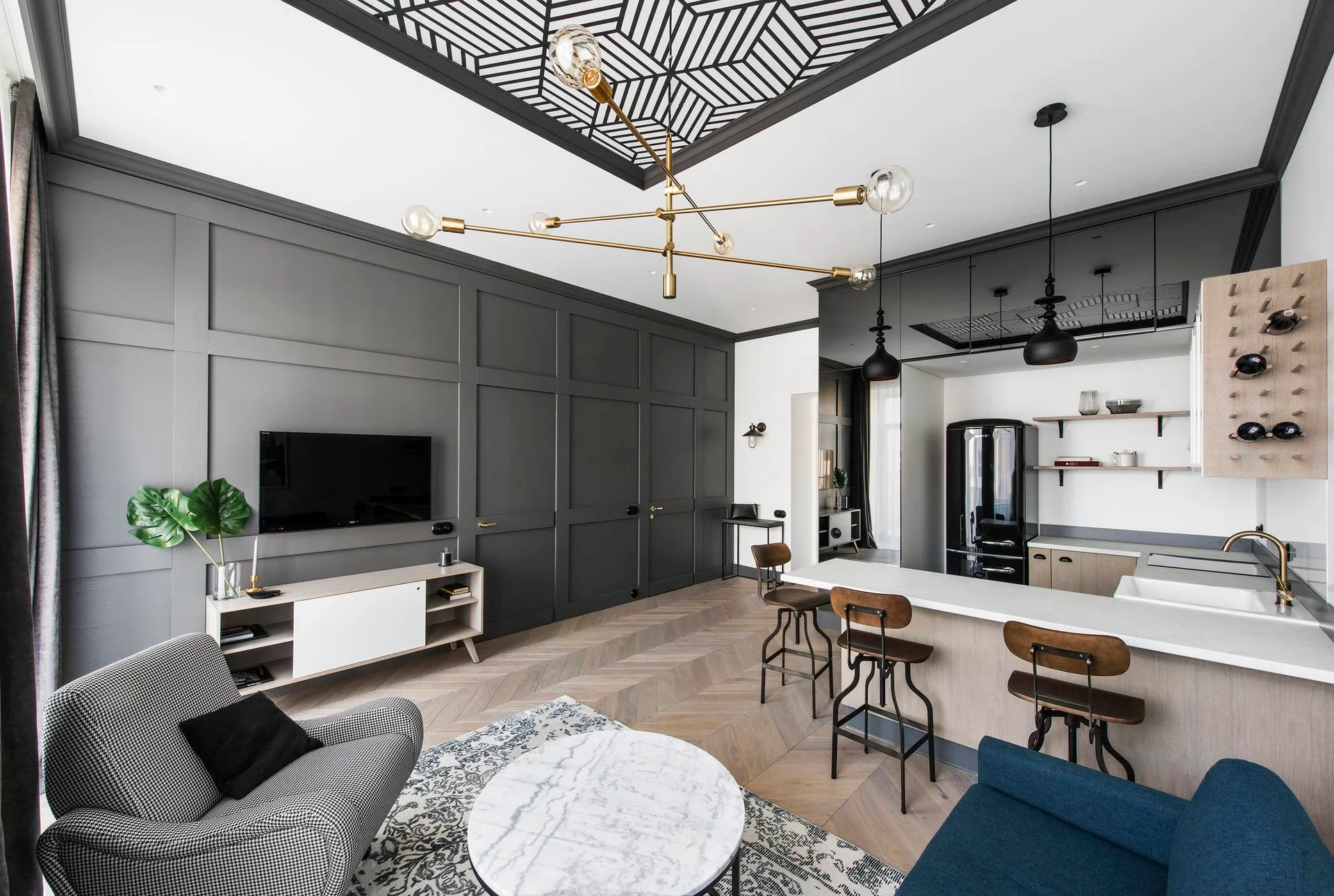 An open-concept living space features a dark gray paneled wall, a kitchen island with stools, a geometric patterned ceiling insert, and chevron wood flooring.