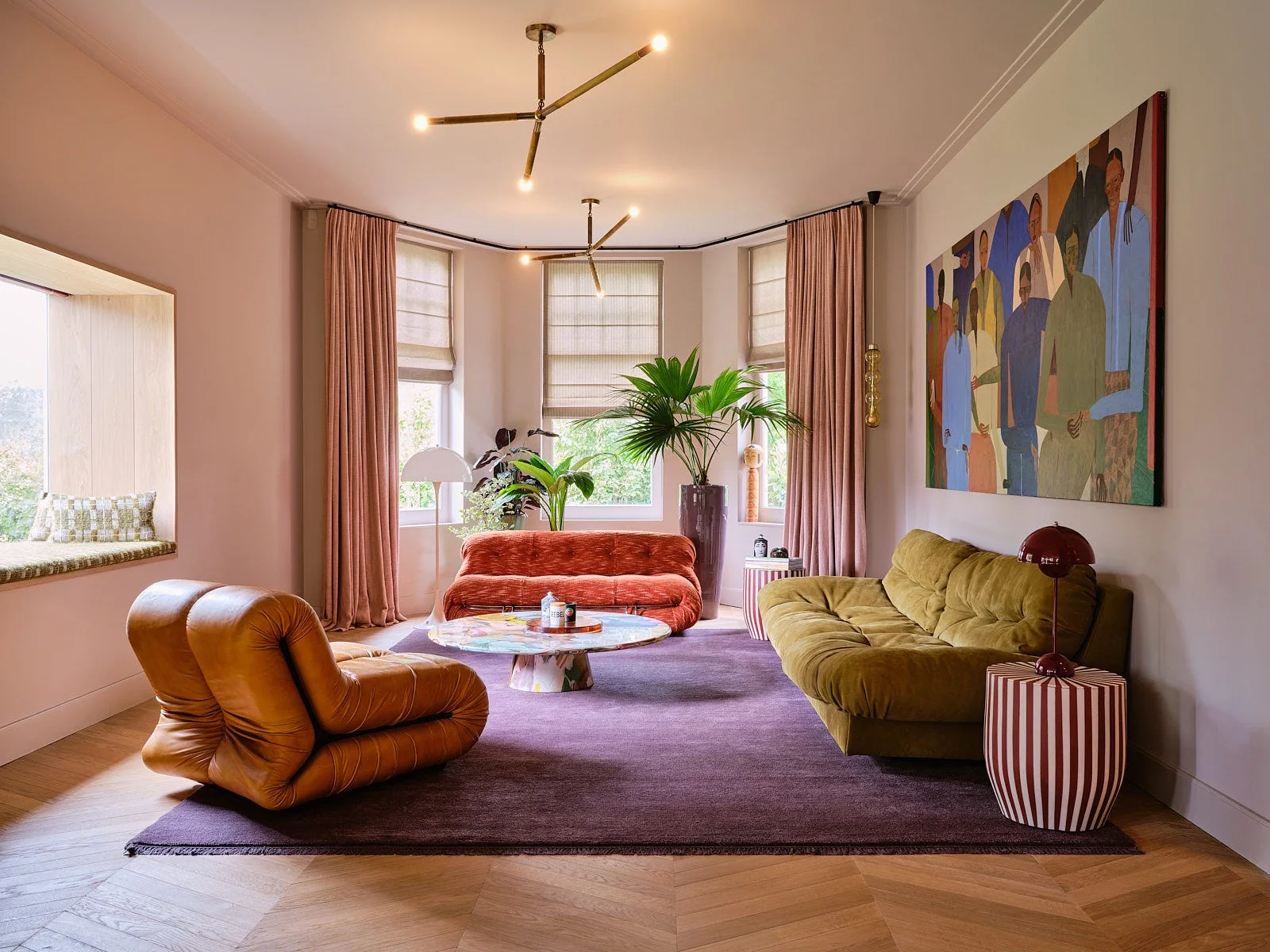 An eclectic living room features a purple area rug, olive green sofa, tan leather lounge chair, and a large colorful painting against pink walls.