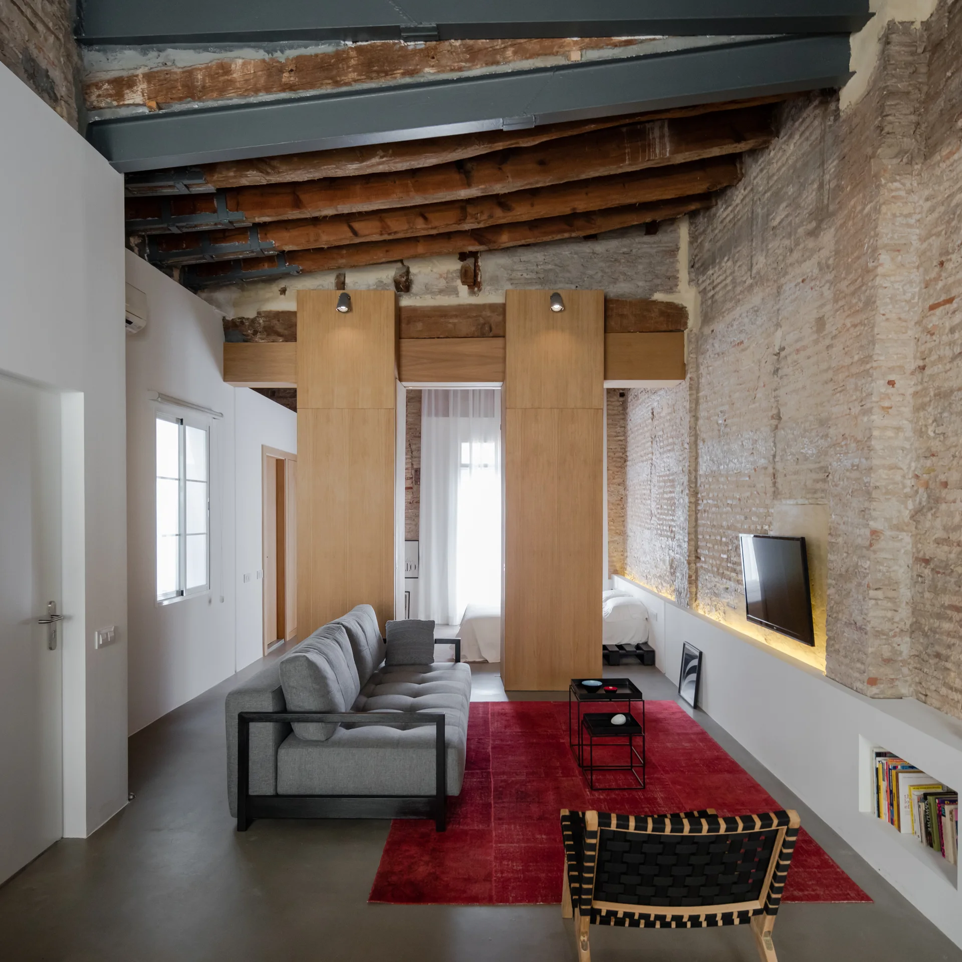 A modern grey sofa sits on a large red rug in an industrial loft featuring exposed brick walls and wooden ceiling beams.