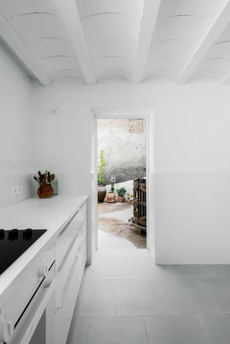 A minimalist white kitchen features flat-front cabinets, a vaulted plaster ceiling, grey floor tiles, and an open doorway leading to a courtyard.