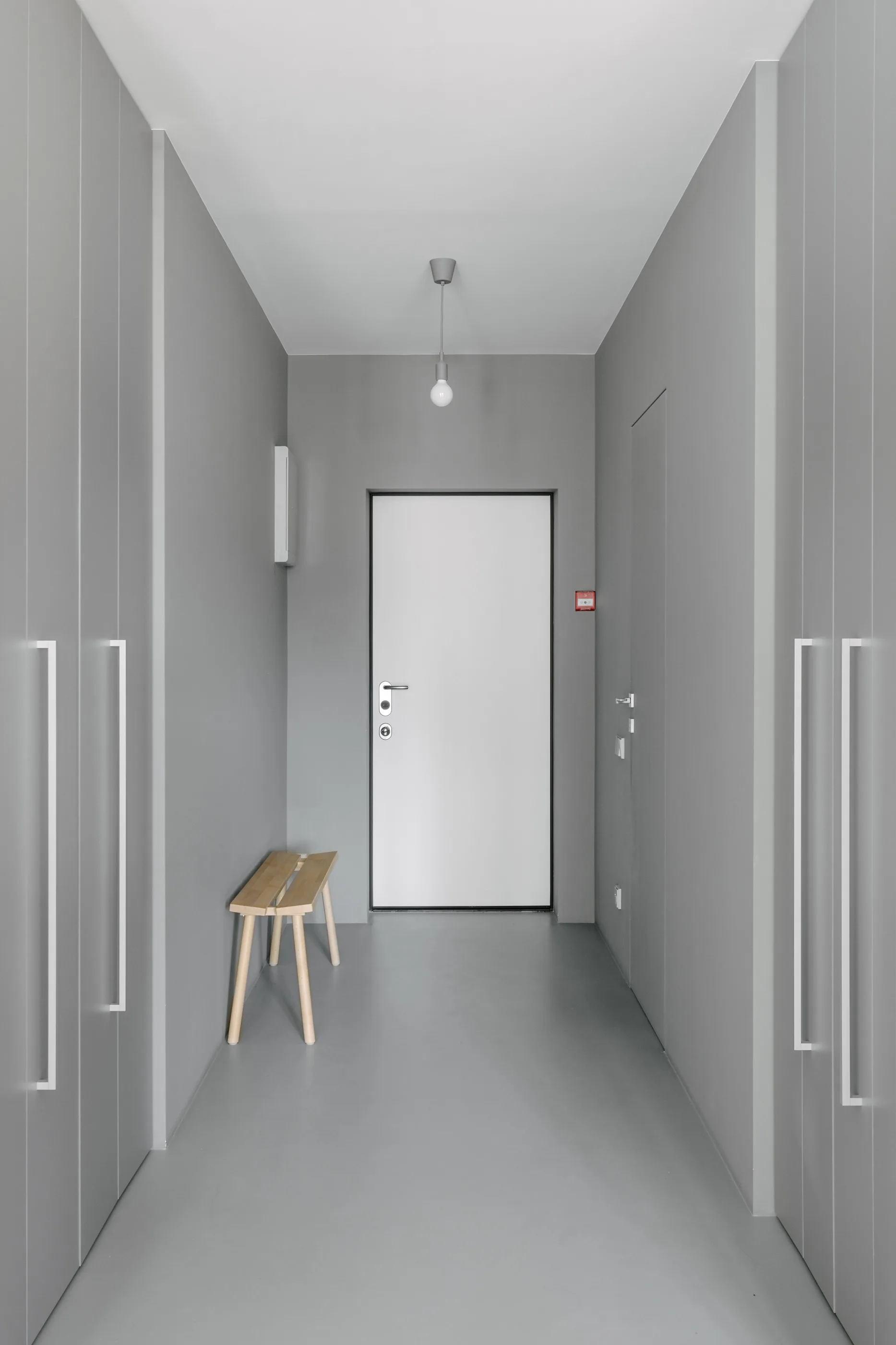A minimalist gray entryway features floor-to-ceiling built-in closets on both sides, a small light wood bench, and a white front door.