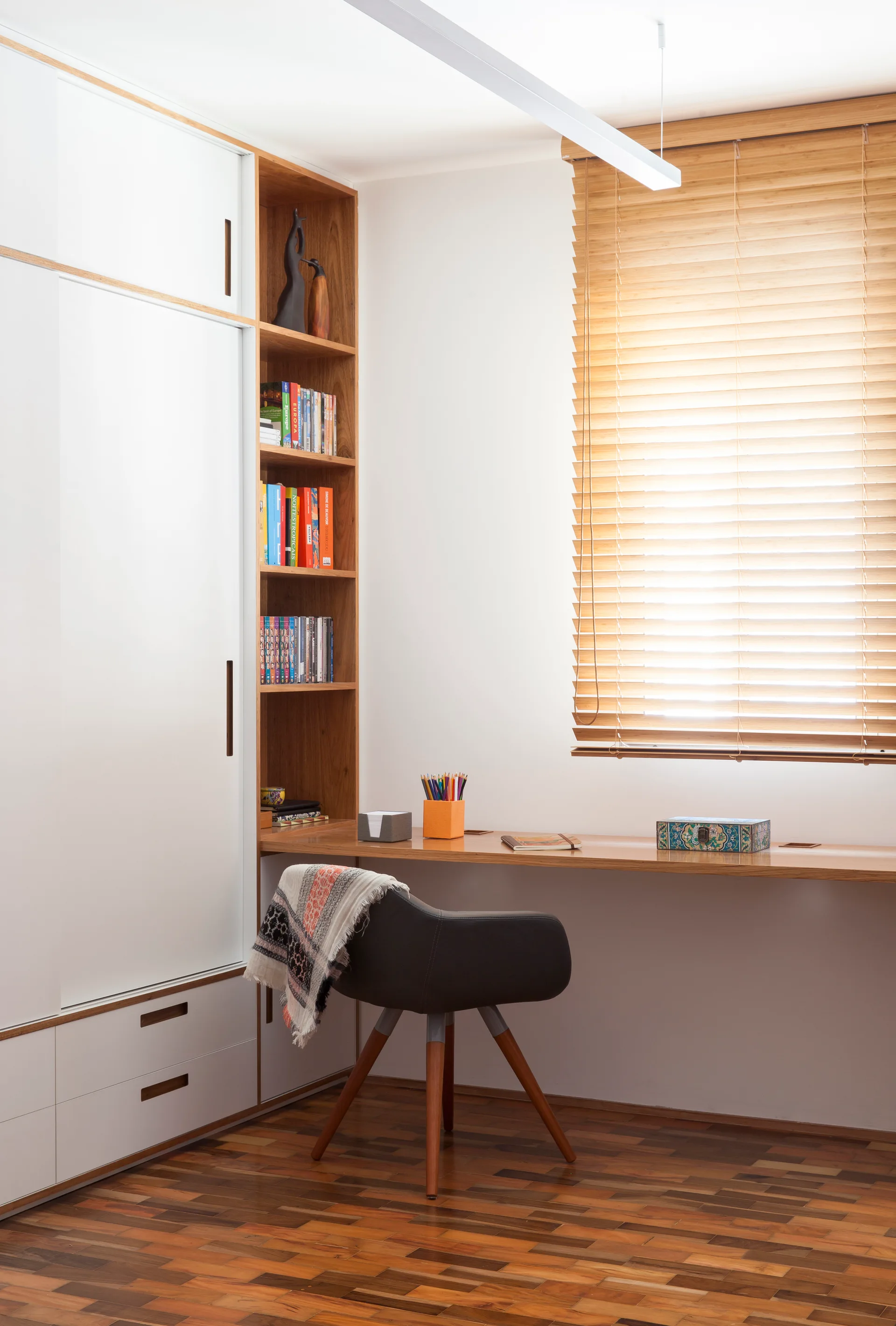 A compact home office workspace featuring a built-in wooden desk integrated with tall white sliding cabinets, open wood shelving, and wooden blinds.