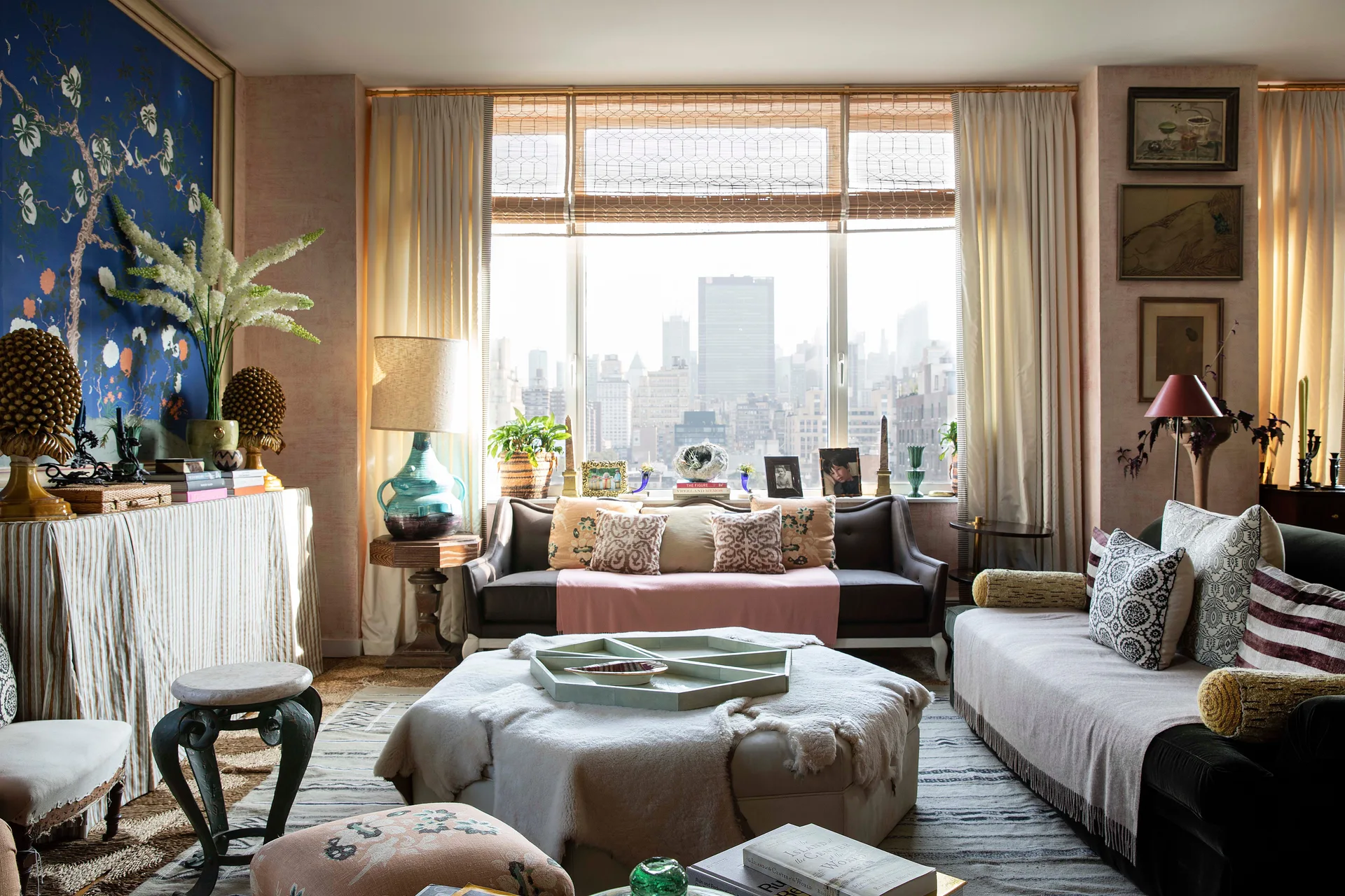 An eclectic living room with a blue floral wall panel, a large window overlooking the city, and a central ottoman topped with a sheepskin rug.