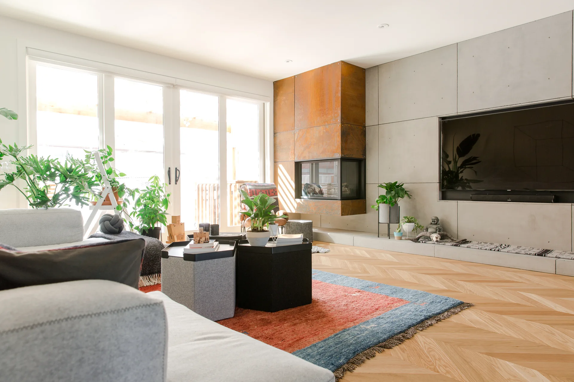 A modern living room features a concrete panel feature wall, a rusted metal fireplace, a grey sofa, and herringbone patterned hardwood floors.