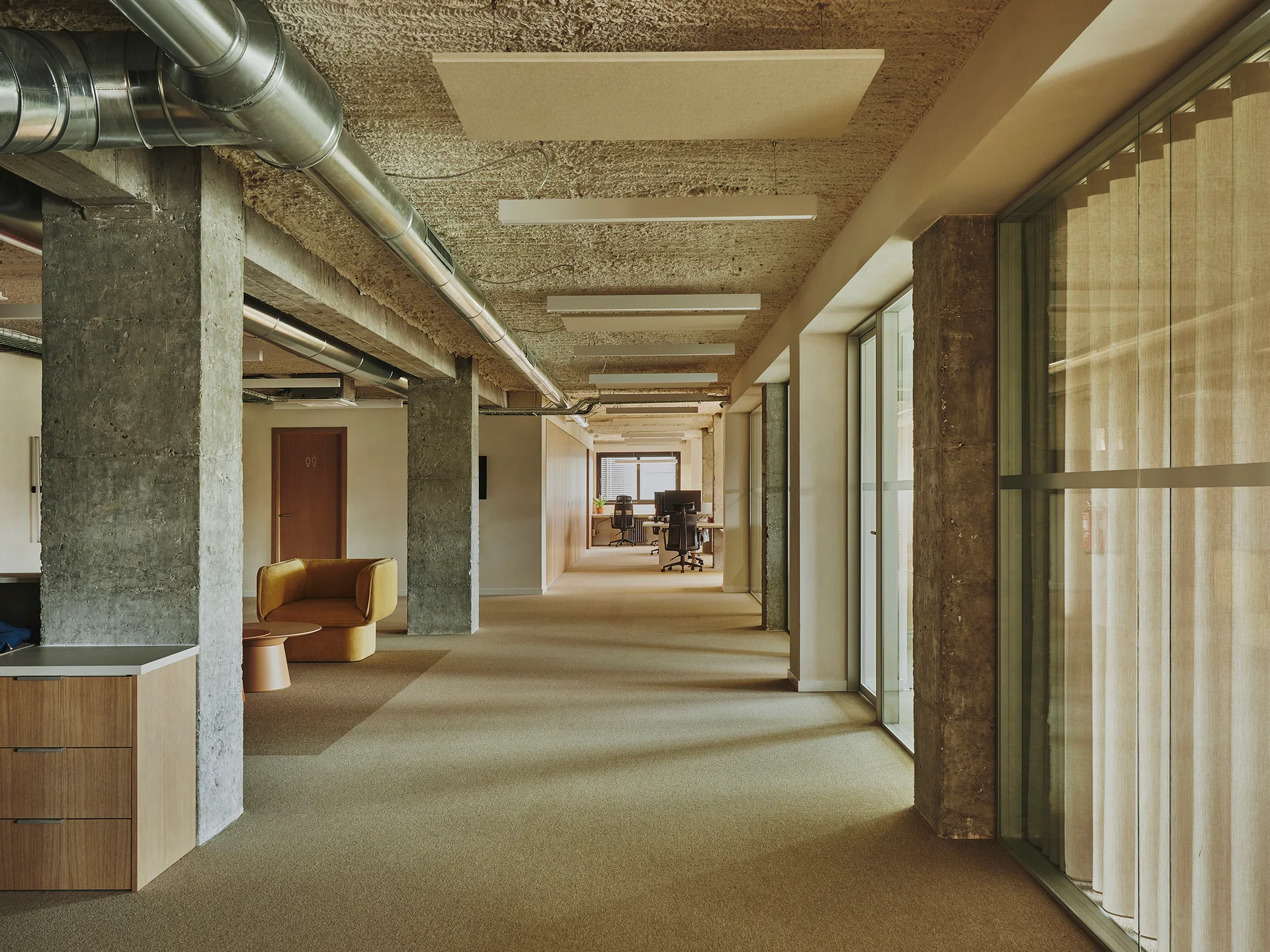 An office hallway featuring exposed concrete pillars, metallic ceiling ductwork, a beige carpeted floor, and a single yellow armchair.