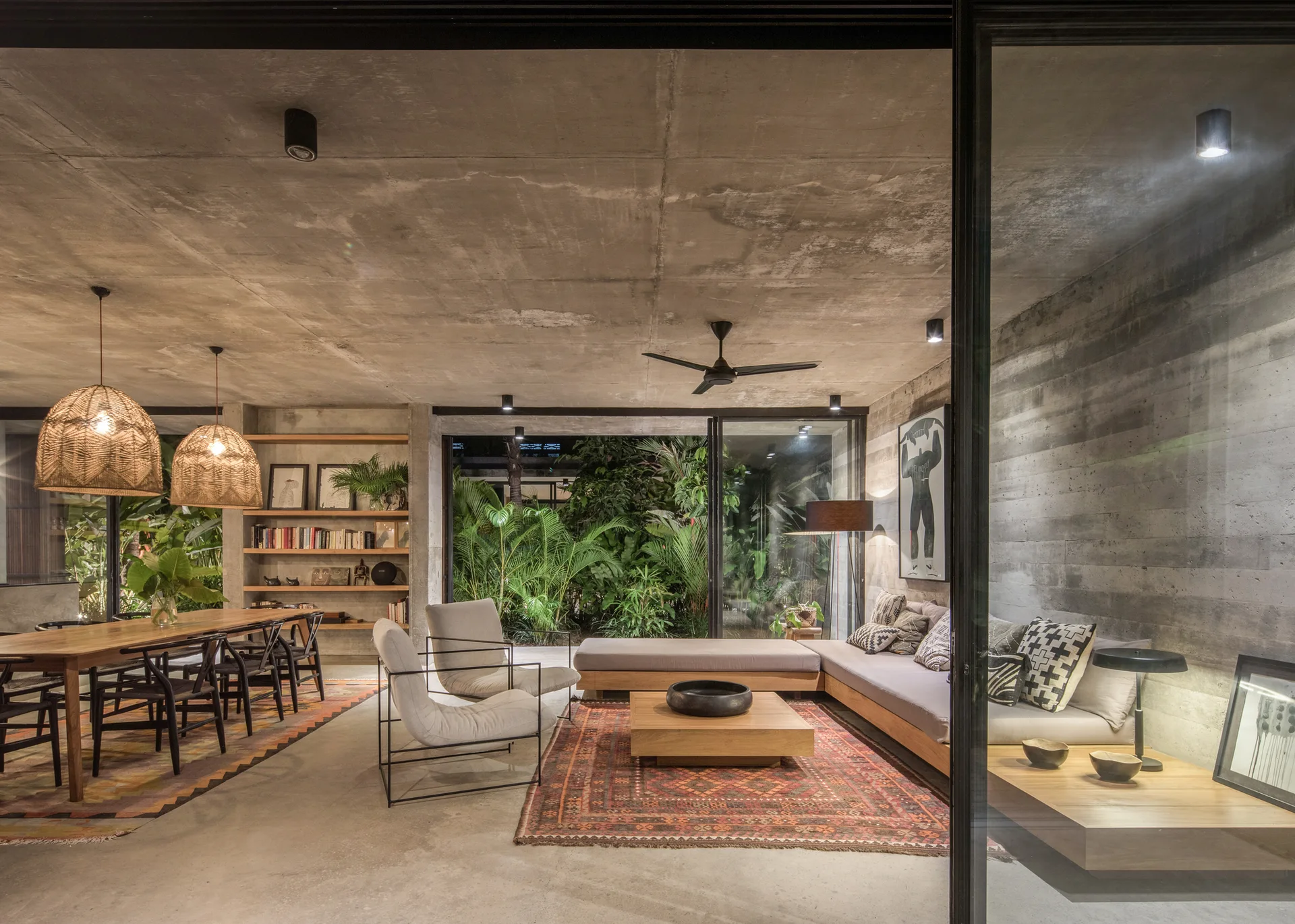 An open-concept living and dining room features raw concrete walls, a low-profile wooden sectional sofa, woven pendant lights, and large glass doors.