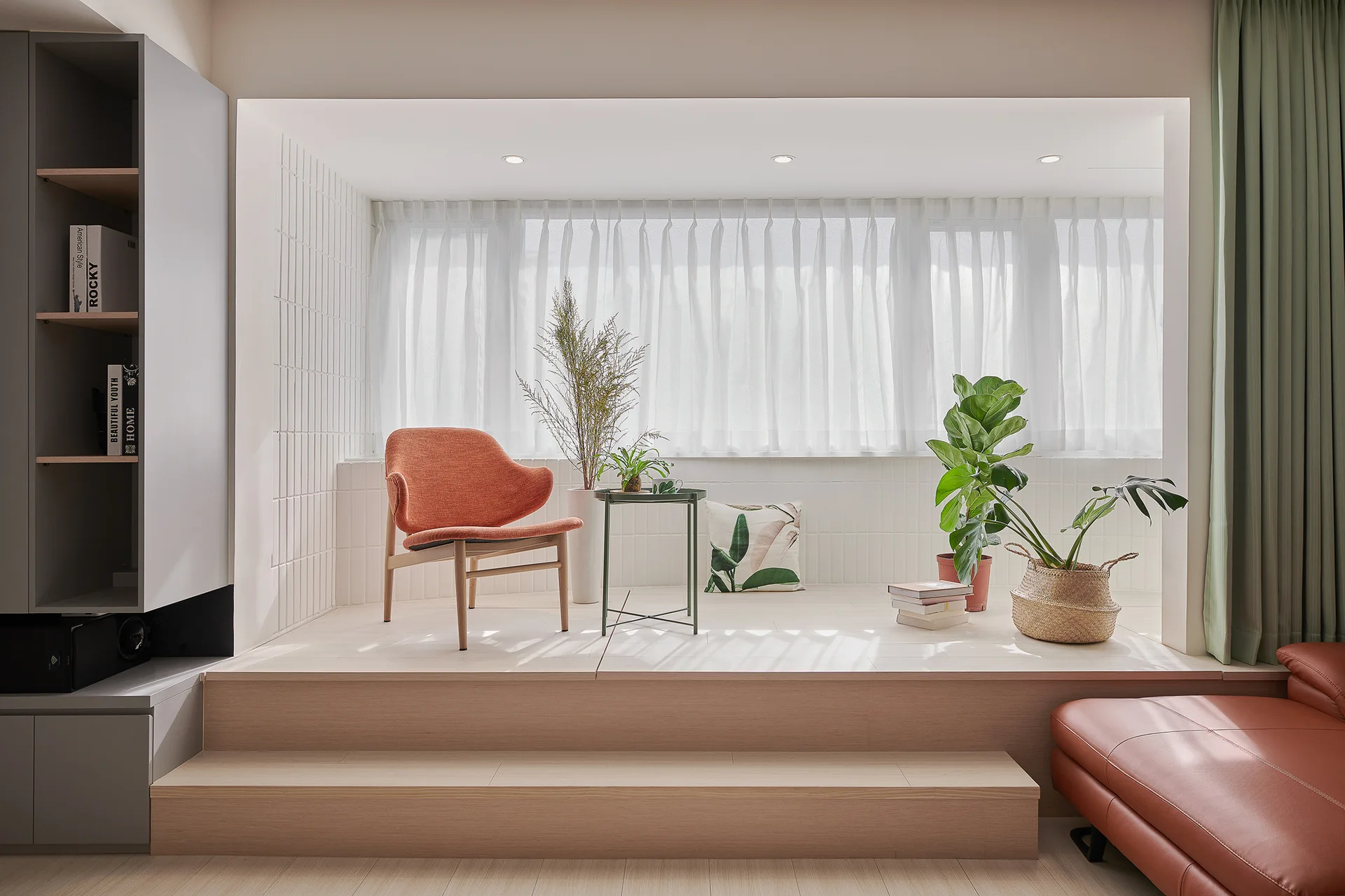 A light wood tiered platform features an orange accent chair, green side table, and potted plants under a window with sheer white curtains.