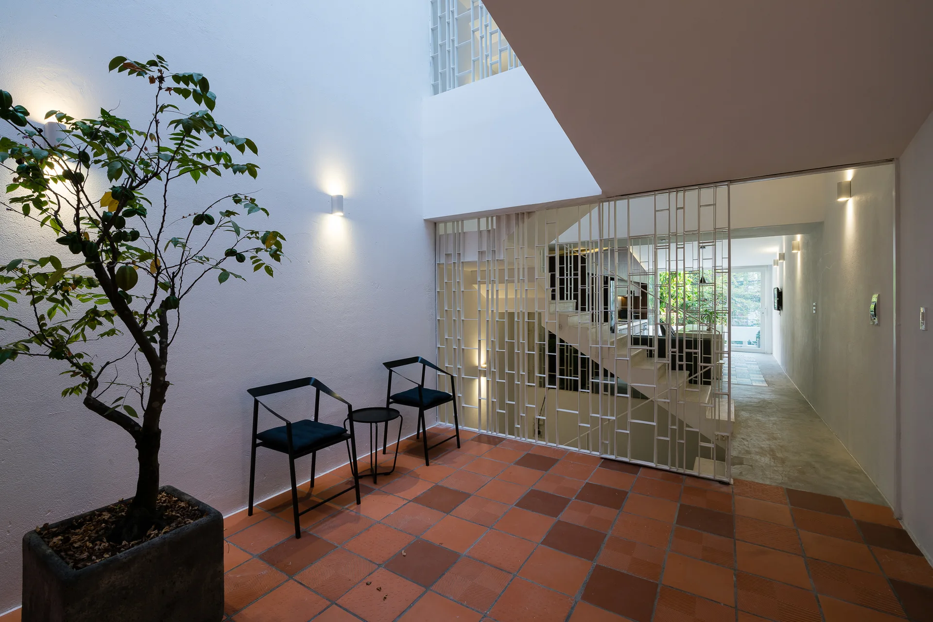 A minimalist entryway features terracotta tile flooring, two modern black accent chairs, a potted tree, and a white geometric metal divider screen.