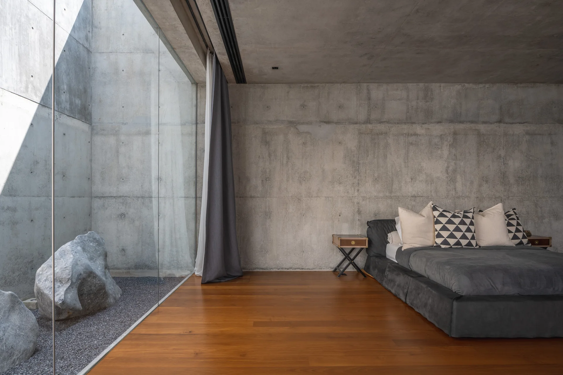A minimalist bedroom features a low grey upholstered bed, warm wooden flooring, and exposed concrete walls next to a glass wall looking out at a rock garden.