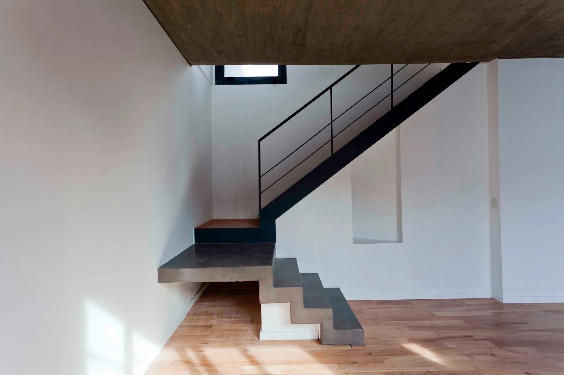 A modern minimalist staircase features gray concrete floating treads, a thin black metal railing, white walls, and light wood flooring.