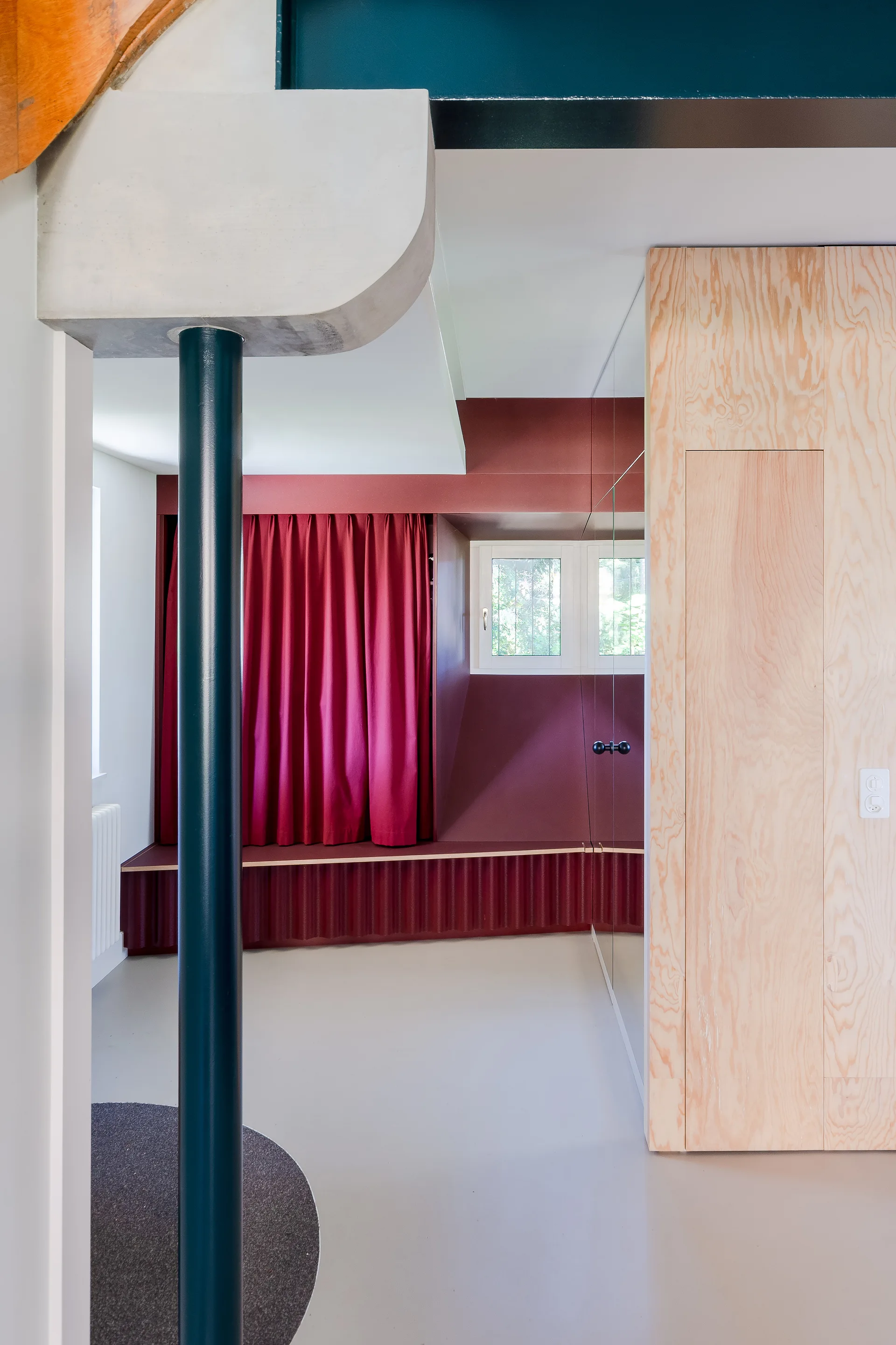 A modernist interior features a burgundy-painted window nook with matching curtains, a corrugated built-in bench, and a light-toned plywood storage cabinet.