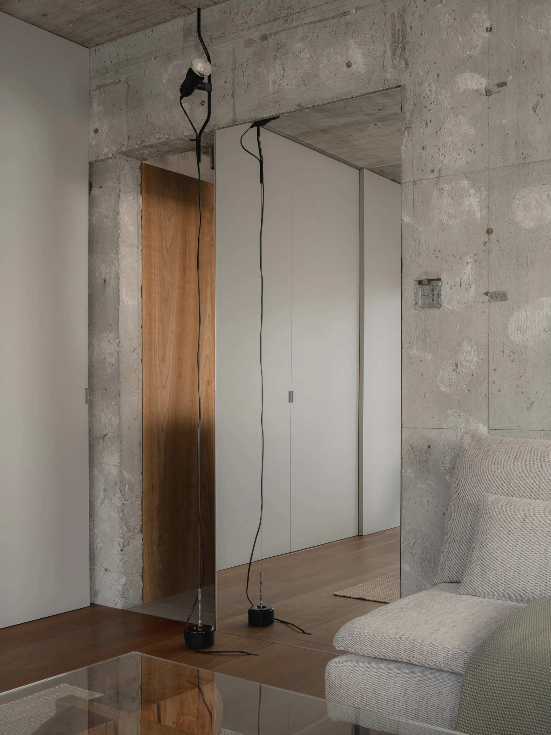 A minimalist living space featuring an exposed concrete wall, a full-height mirror, a black cable pendant light, and white integrated cabinetry on wood floors.