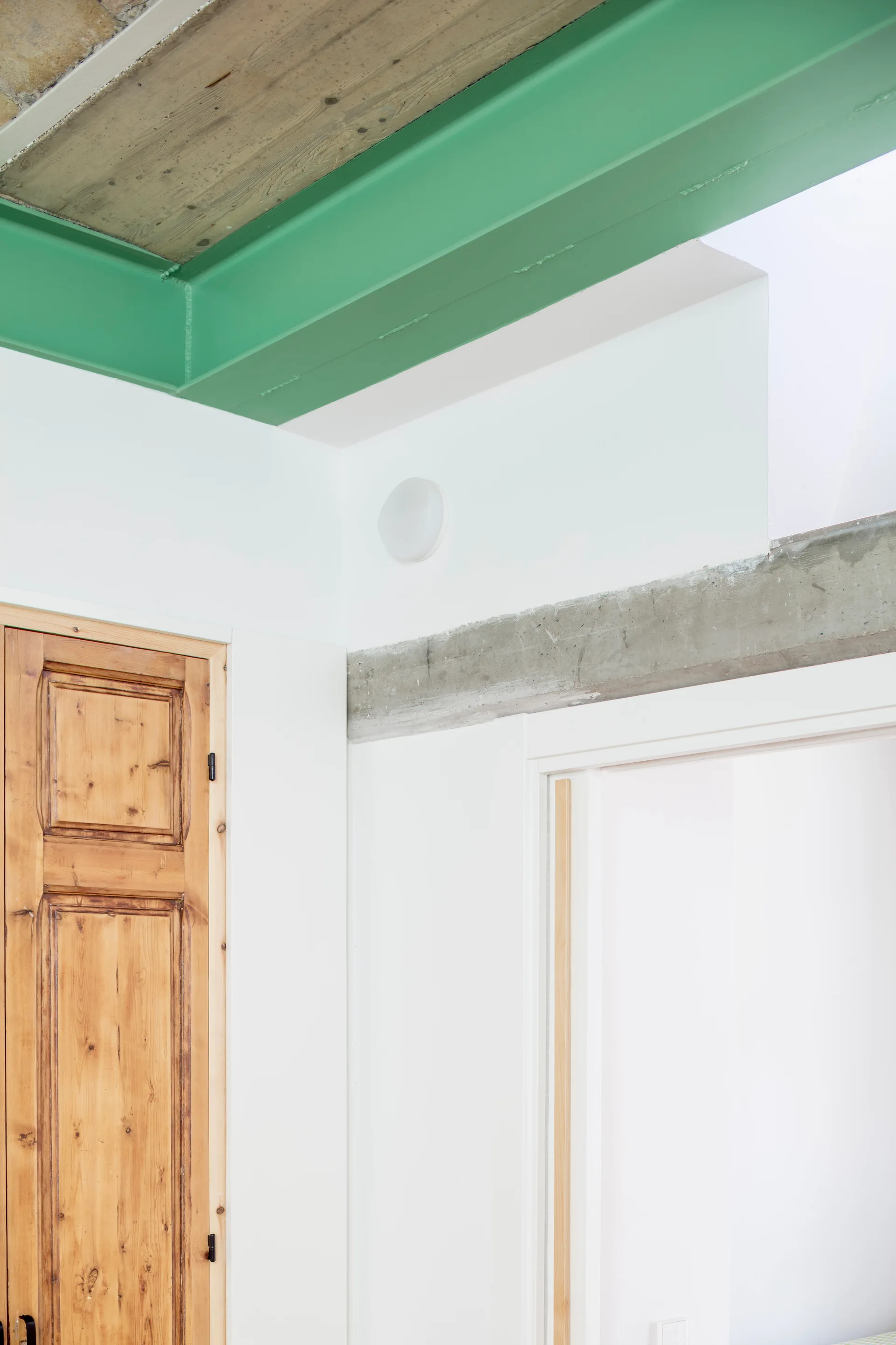 An interior corner showing a bright green painted structural beam, an exposed gray concrete lintel, and a natural wood paneled door.