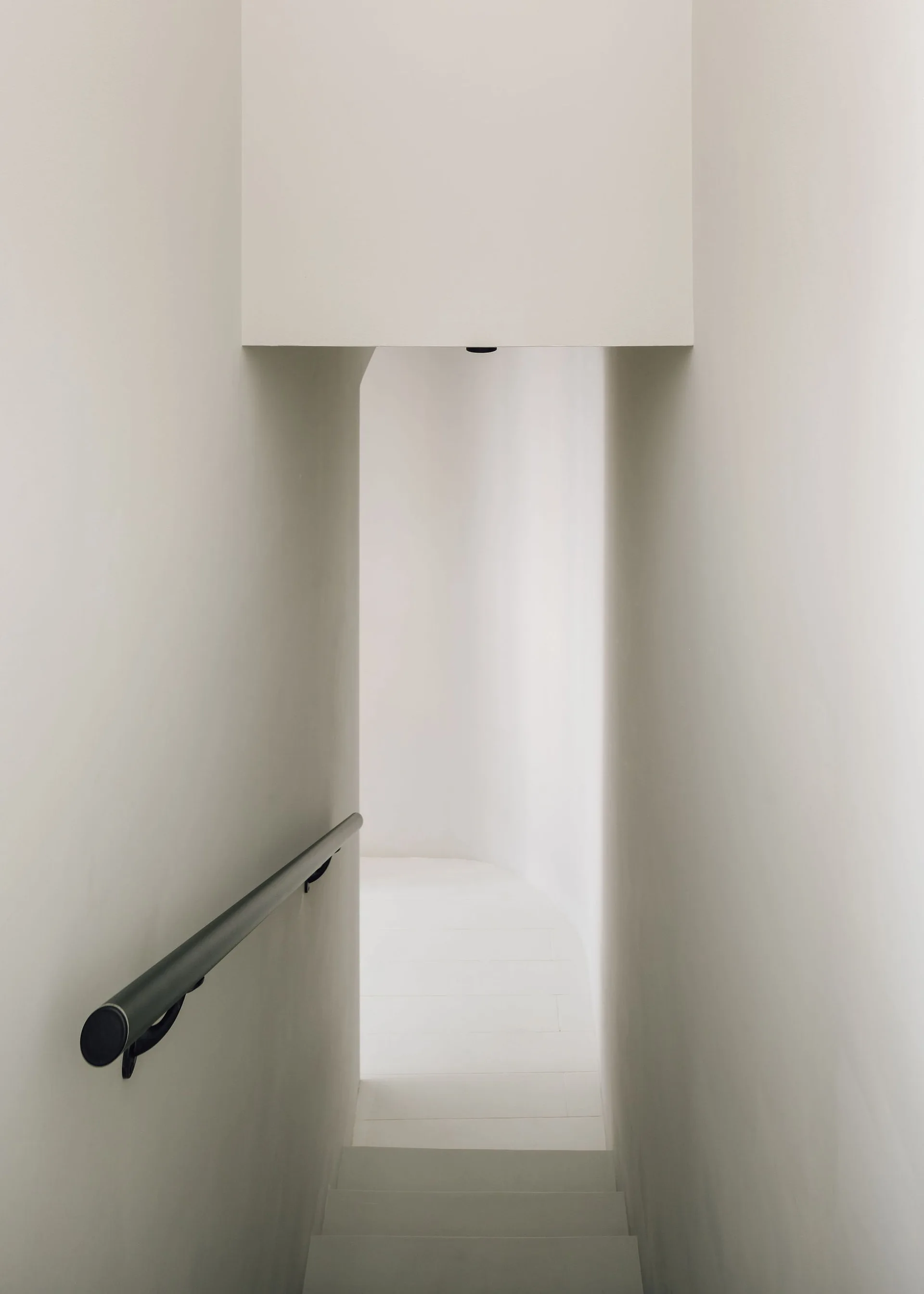 A minimalist white plaster staircase descends between clean matte walls with a dark metal handrail leading toward a curved passage.