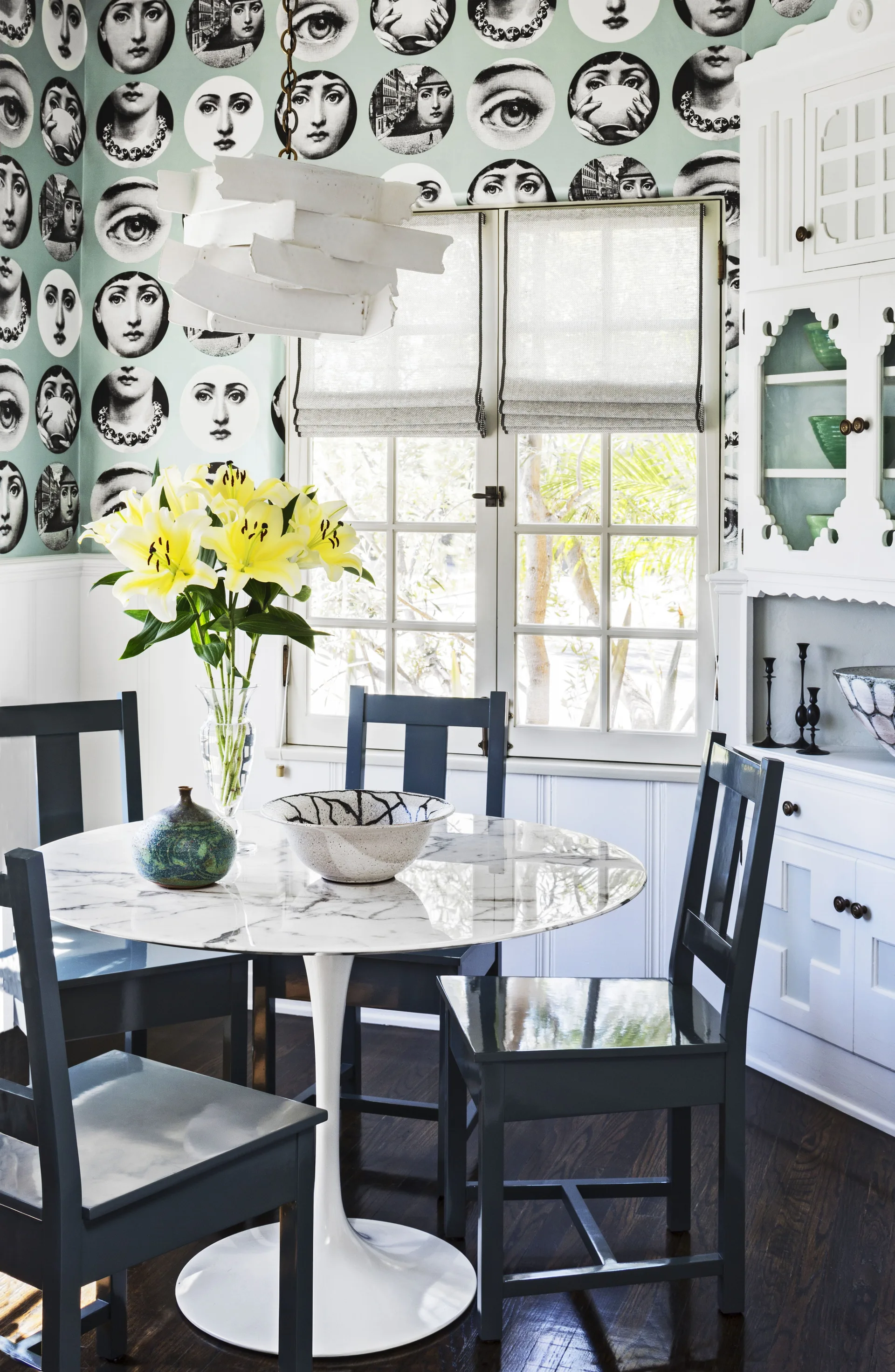 A dining room features a round marble tulip table with dark wood chairs set against mint wallpaper with a face motif and white wainscoting.