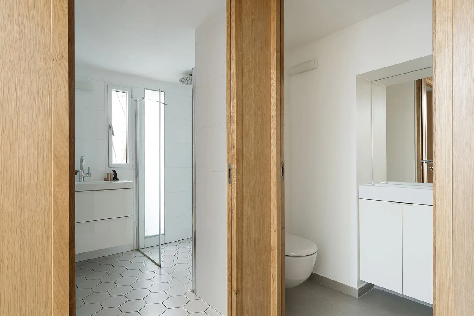 A minimalist split bathroom featuring oak-framed doorways, white floating vanities, and hexagonal floor tiles.