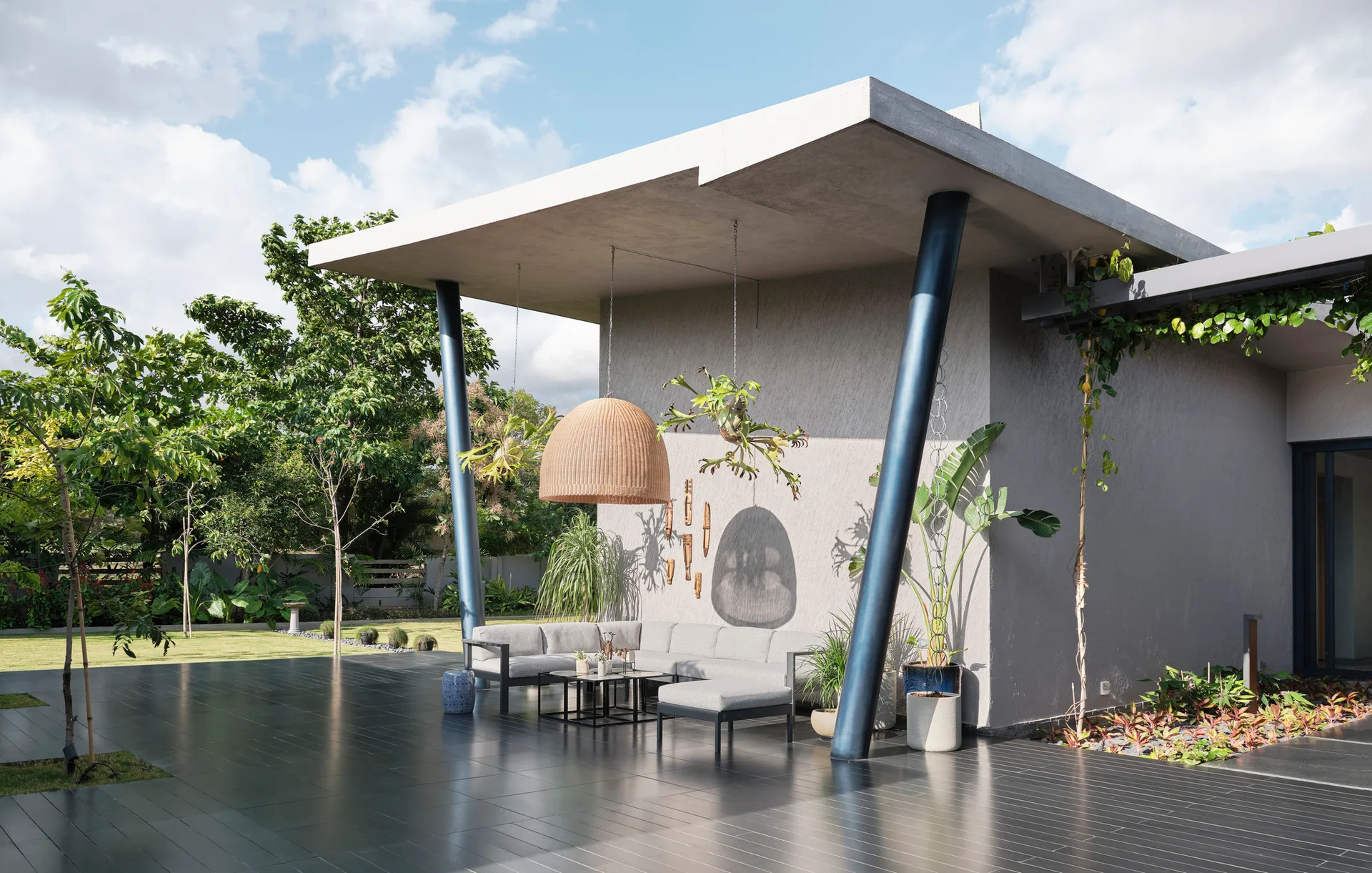 A modern covered concrete patio with a gray sectional sofa, a large wicker pendant light, and slanted blue metal columns overlooking a garden.