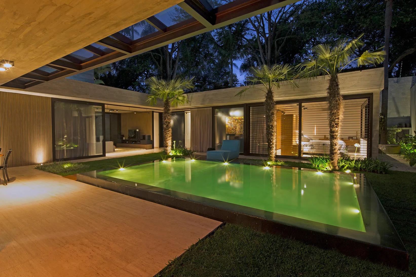 An illuminated green swimming pool sits in a modern concrete courtyard surrounded by palm trees, a stone patio, and glass sliding doors.