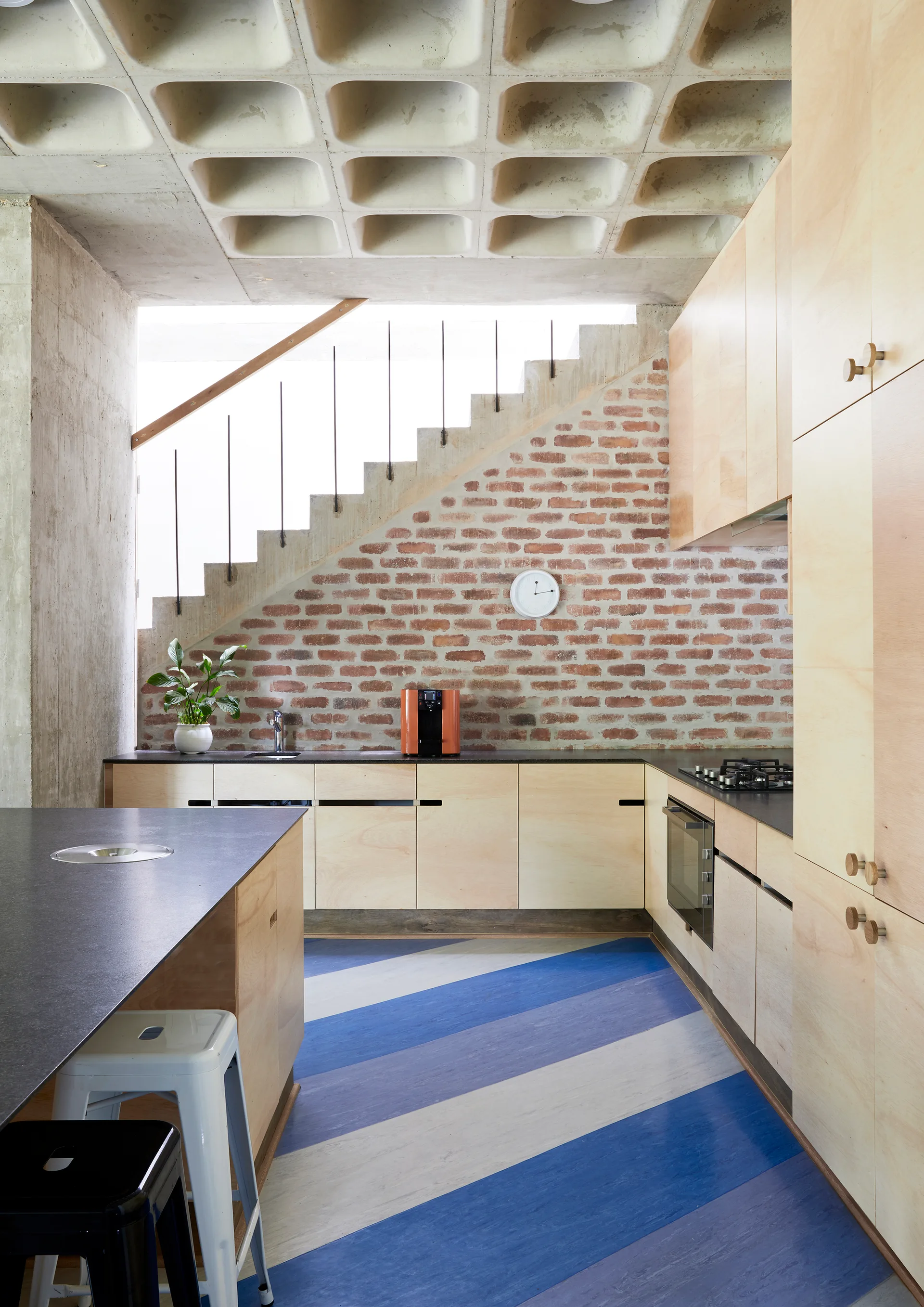 A modernist kitchen features light wood cabinets, an exposed brick wall, a concrete waffle ceiling, and a bold blue and white striped floor.