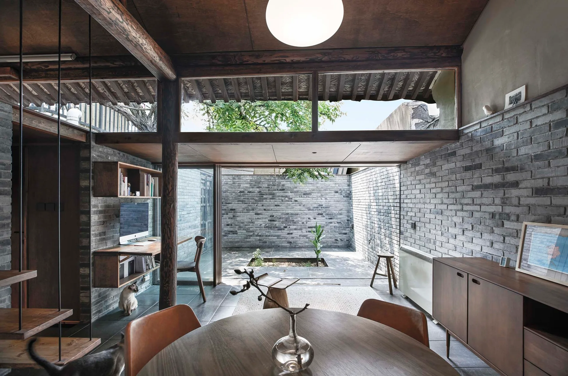 A dining room featuring a round wooden table, grey brick walls, and a large glass door opening onto a private stone courtyard.