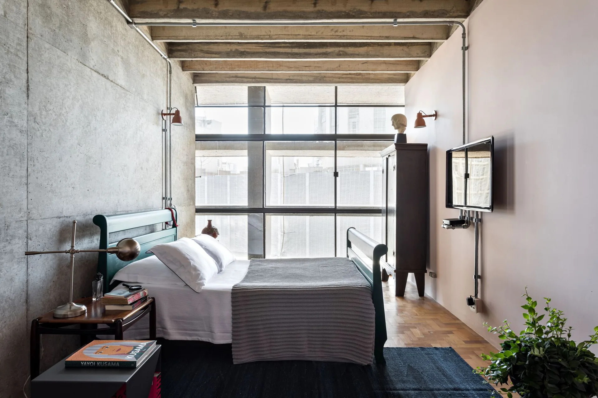An industrial loft bedroom features an exposed concrete wall and ceiling, a green wooden bed, and a large metal-framed window.