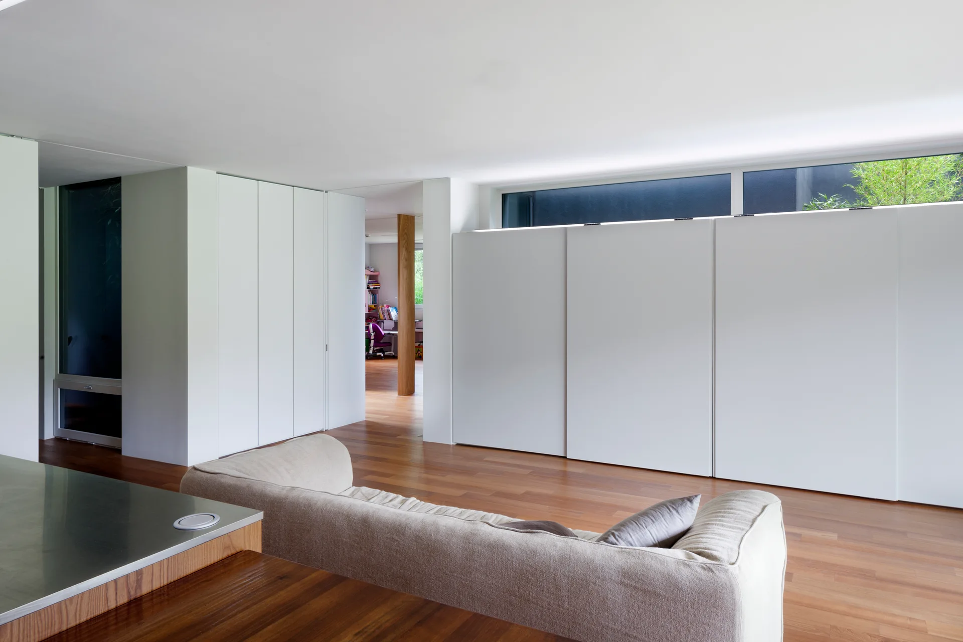 A minimalist living room features a beige sofa, hardwood floors, large white built-in storage cabinets, and a long horizontal clerestory window.