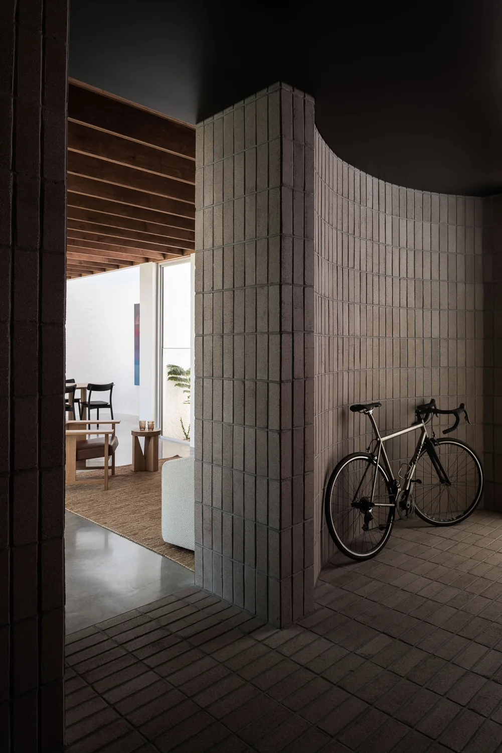 A bicycle stands against a curved grey brick wall in an entryway that transitions to a living room with exposed wood ceiling beams.