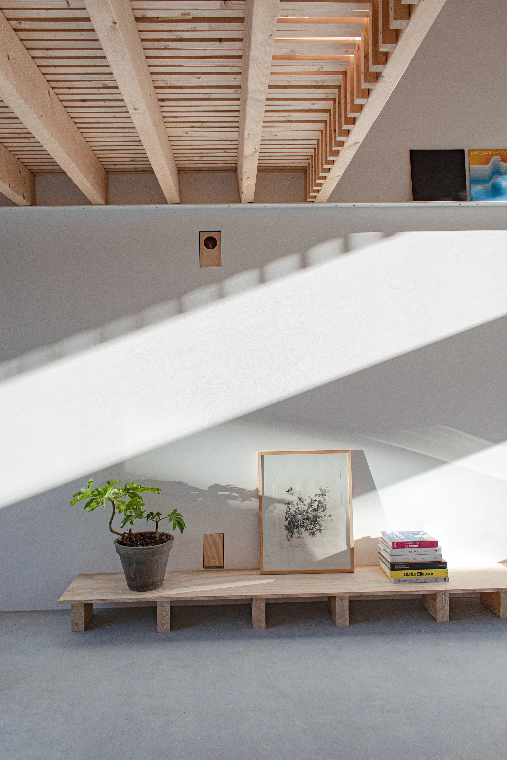 A low plywood bench with a potted plant, framed art, and stacked books sits against a white wall under exposed timber ceiling beams.