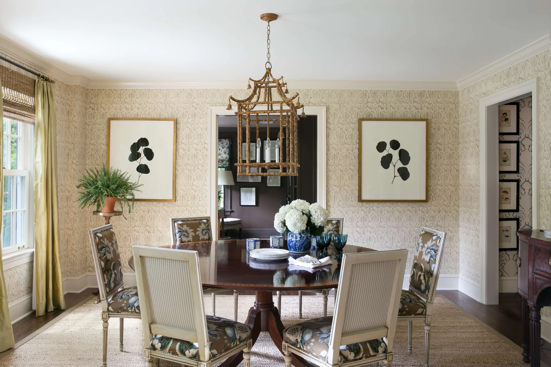 A traditional dining room features a round wood table, floral chairs, a bamboo lantern chandelier, and patterned wallpaper with framed botanical prints.