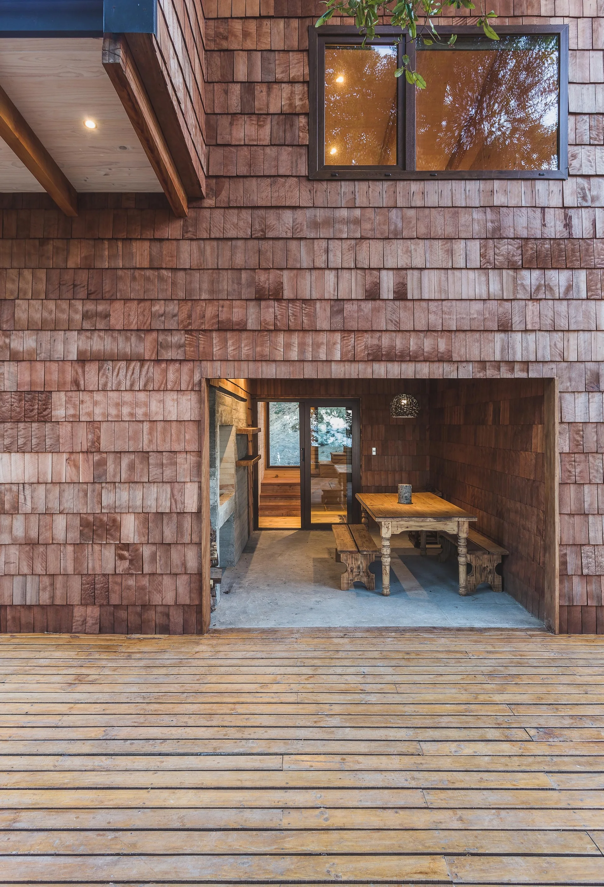 A covered outdoor patio with a wooden dining table and benches, nestled into a recess of a brown wood shingle exterior wall next to a deck.