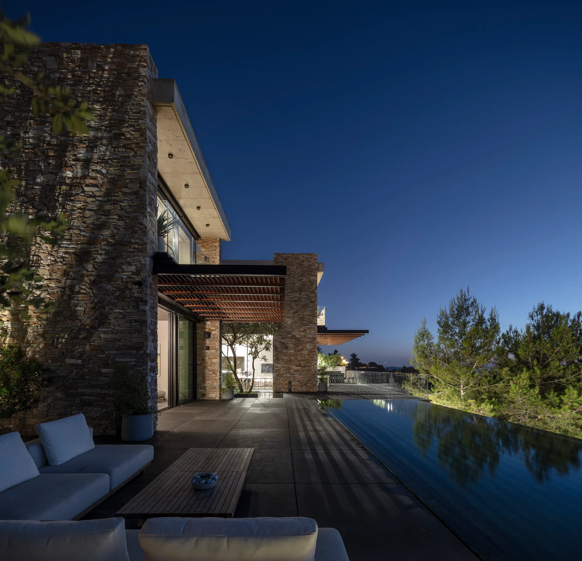 A dark concrete patio features a low-profile sectional sofa, a slatted wood coffee table, and an infinity pool next to a stacked stone house at night.