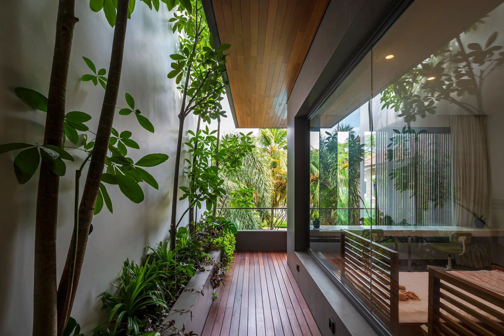 A narrow modern balcony features warm wood decking, matching ceiling planks, a built-in concrete planter with green plants, and a glass wall.