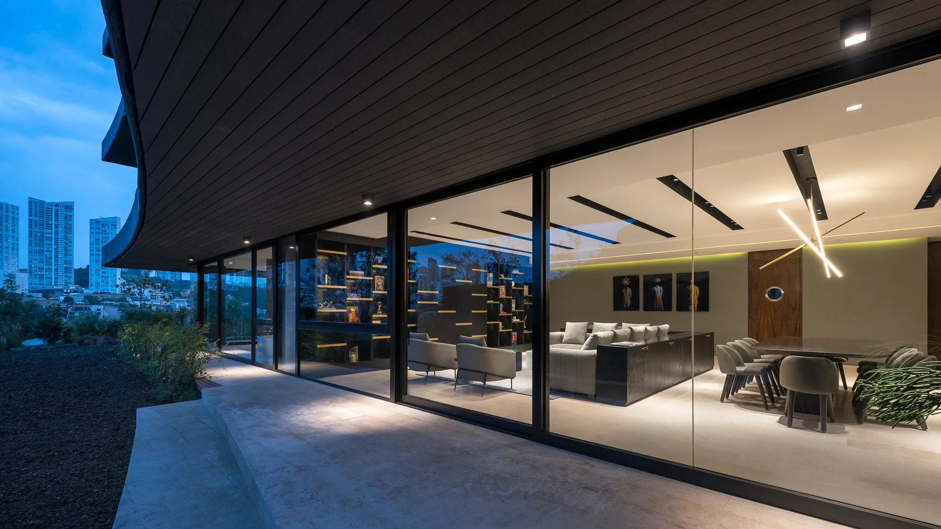 A twilight view from a stone terrace looking through a large glass sliding wall into an illuminated modern living and dining room with a grey sectional sofa.