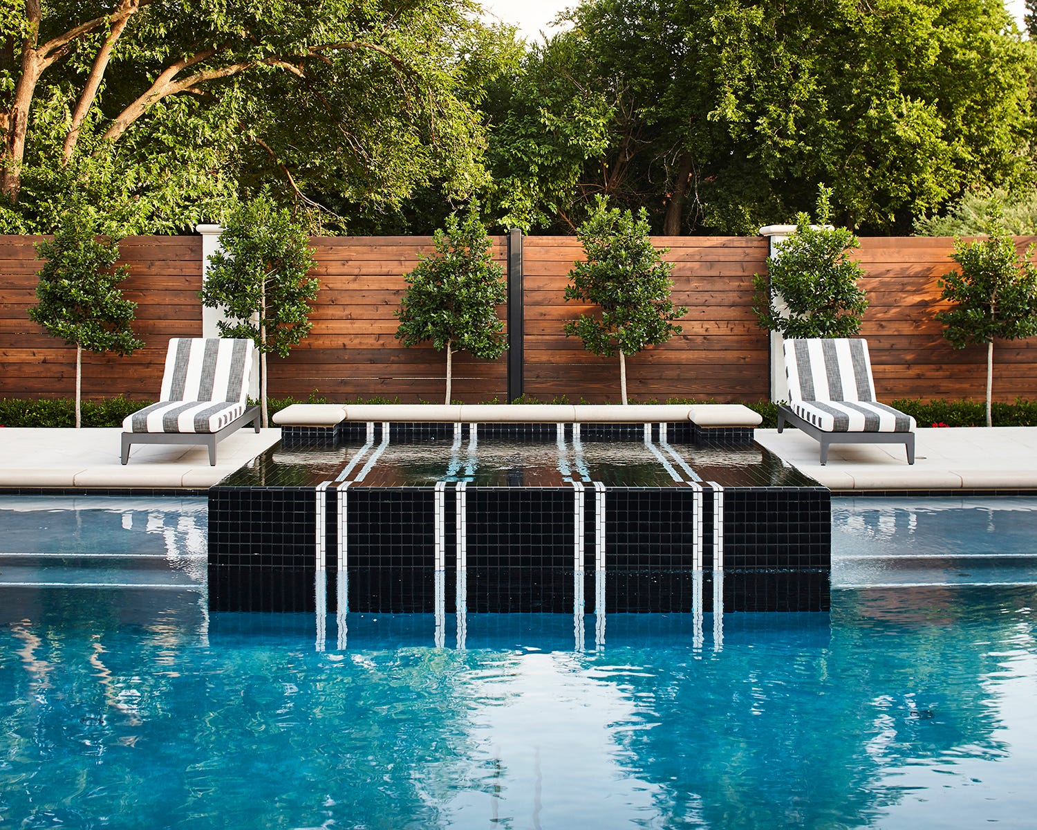A backyard swimming pool features a raised black-tiled spa with white stripes, flanked by two striped lounge chairs against a horizontal wood fence.