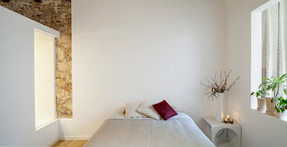 A minimalist bedroom featuring a bed with neutral bedding, a white wall, a vertical strip of exposed stone, and a recessed window ledge with potted plants.