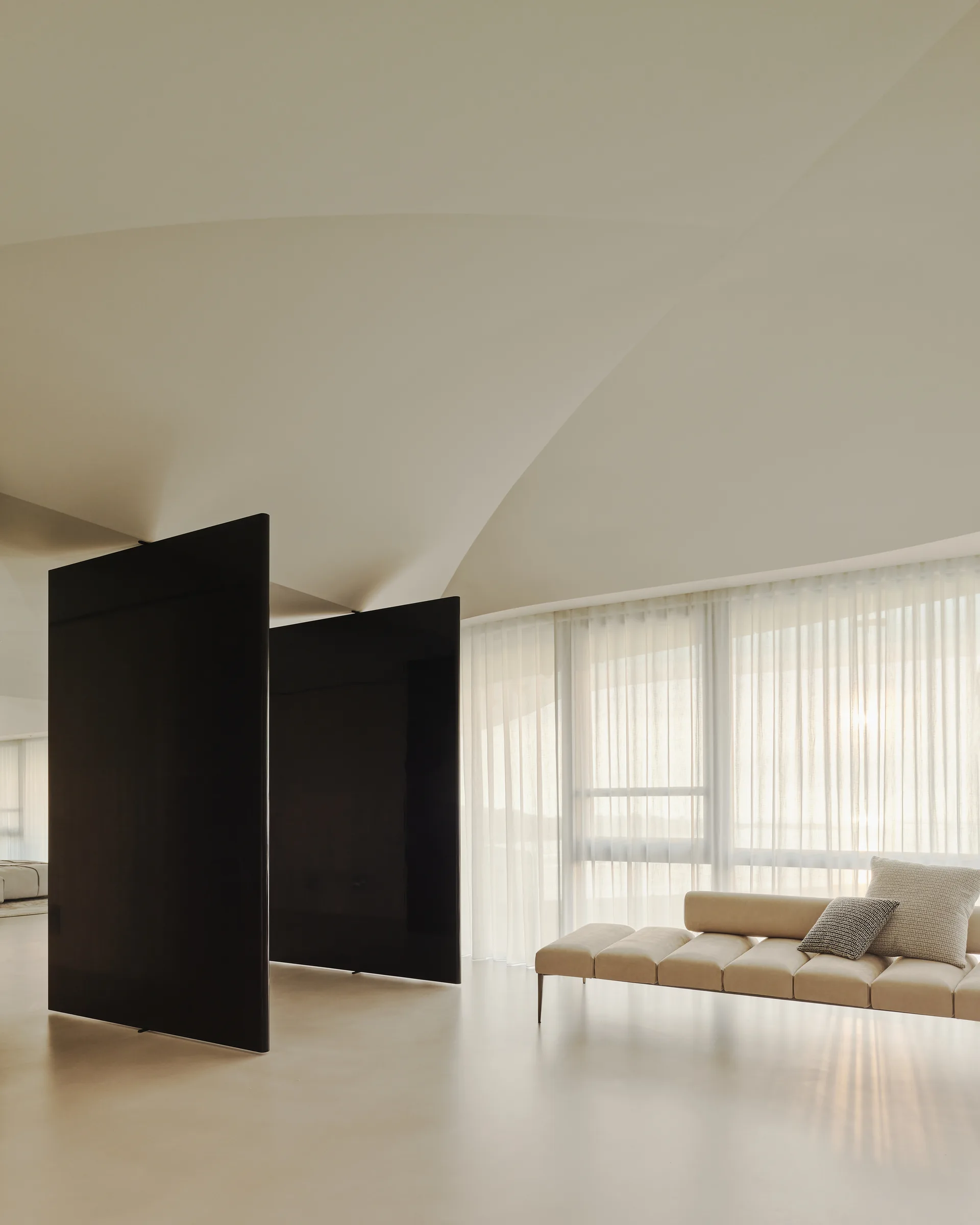 A minimalist living room features a beige upholstered daybed, sheer curtains, tall black room divider panels, and a curved architectural ceiling.