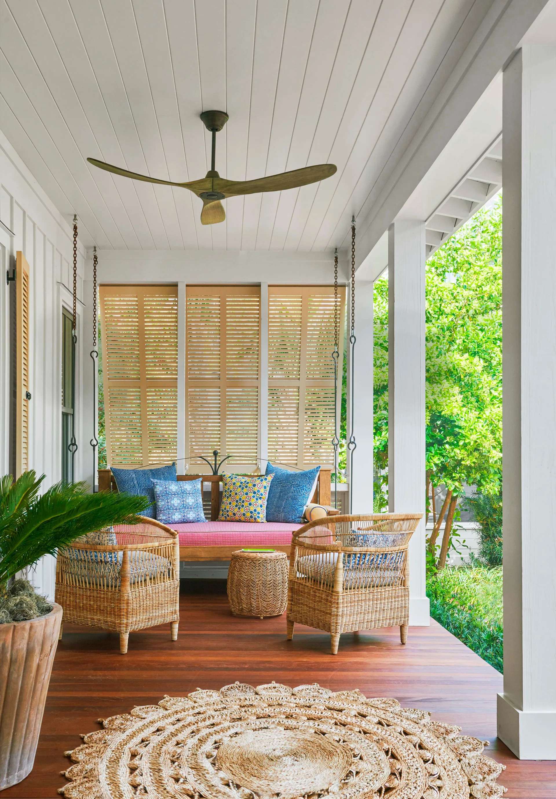 A covered porch featuring a suspended wooden swing, two rattan armchairs, a round braided jute rug, and wooden shutters in the background.