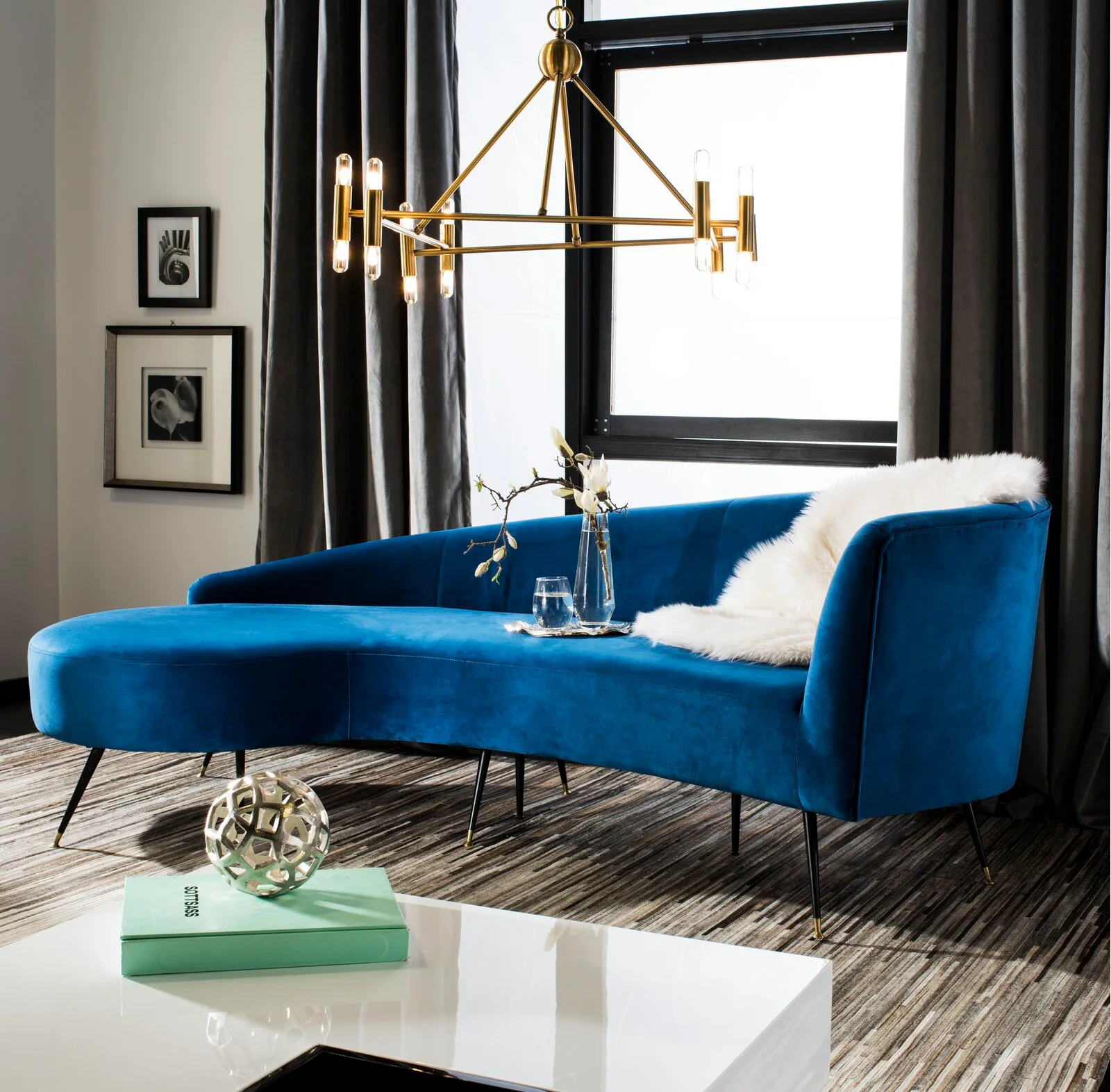 A curved royal blue velvet sofa rests on a striped grey rug under a modern brass chandelier in front of dark grey curtains.