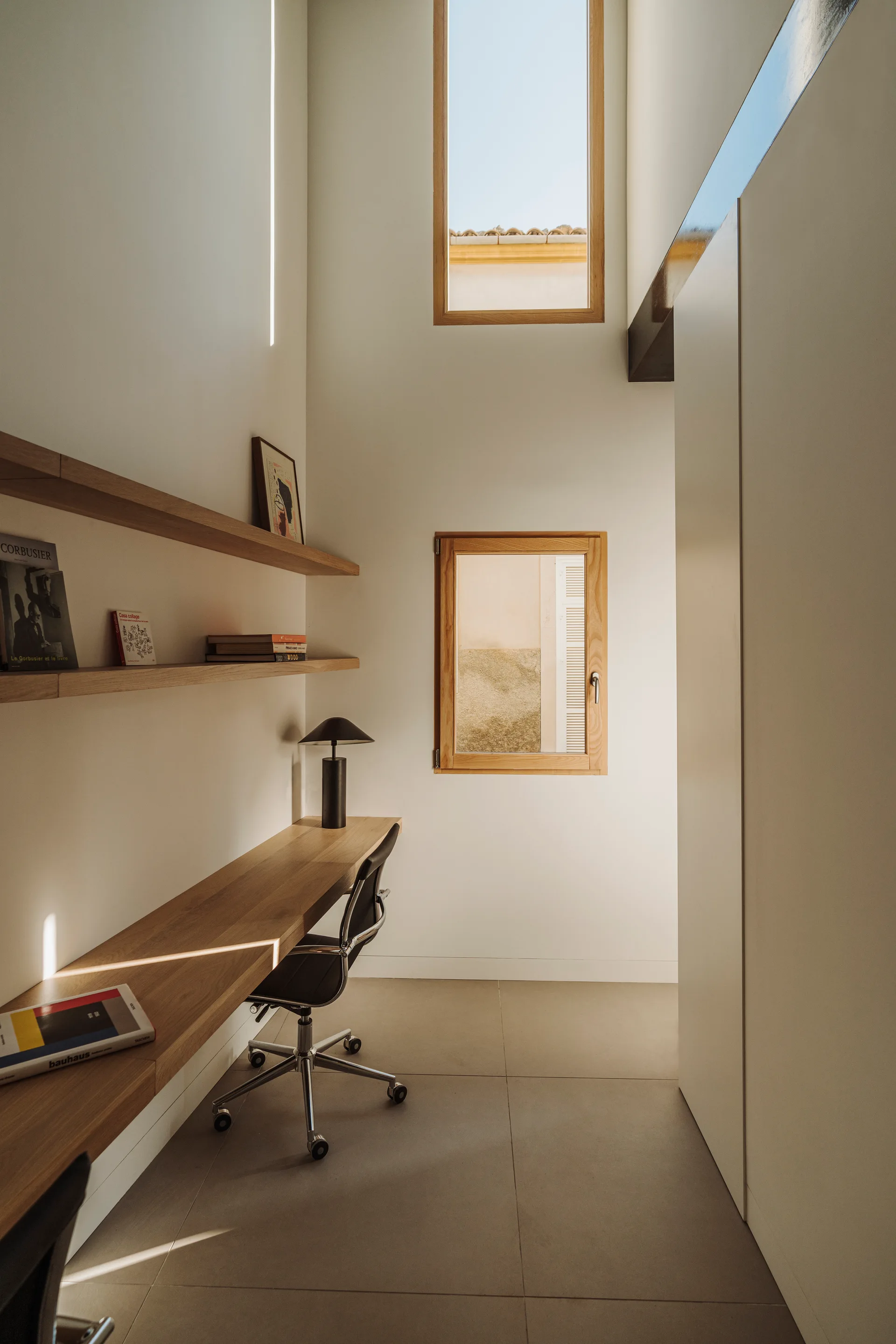 A minimalist home office with a wall-mounted wooden desk, black swivel chair, floating shelves, and two stacked windows set in white walls.