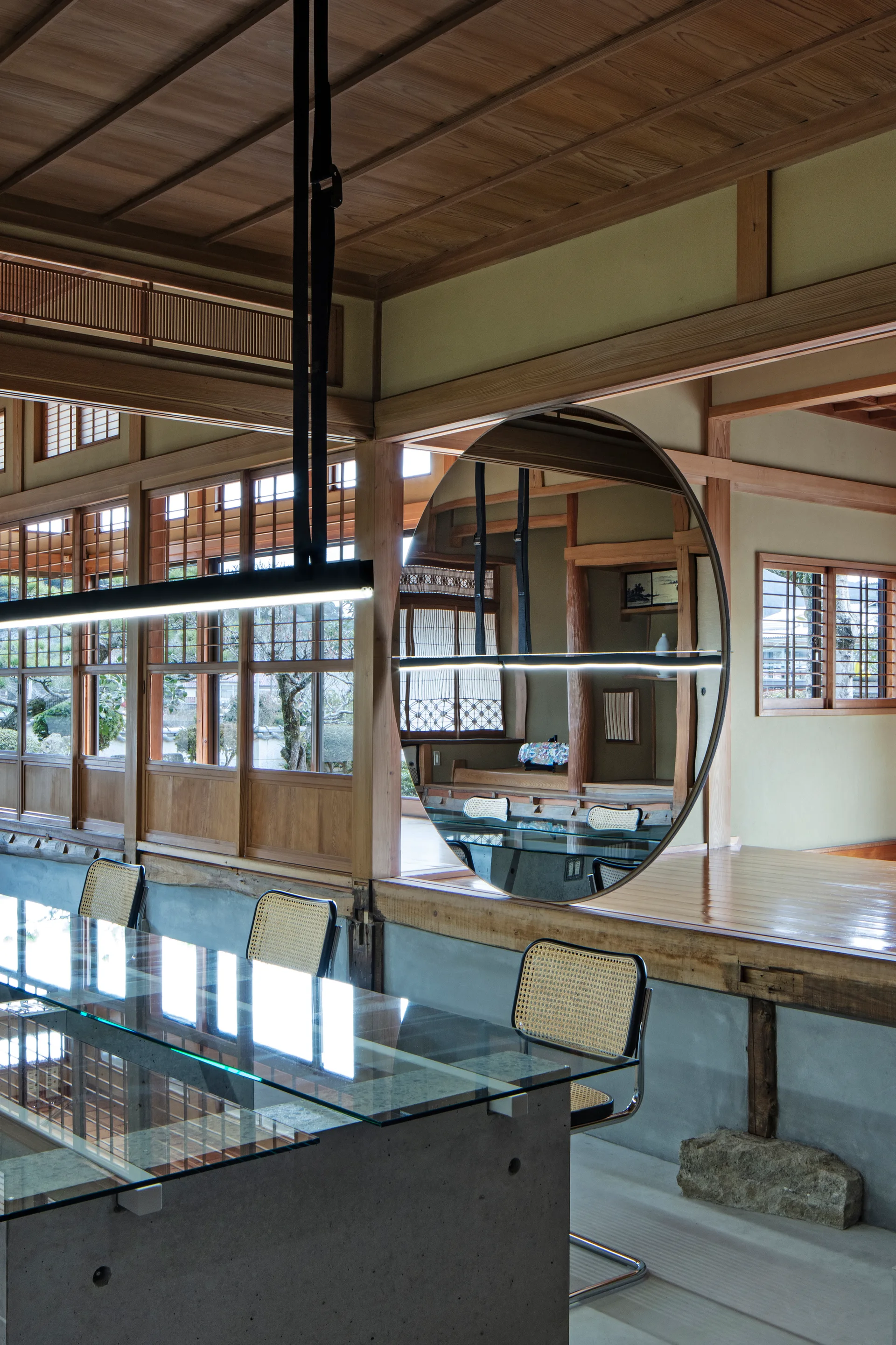 A contemporary glass table with concrete bases stands in a timber-framed room with a large round mirror reflecting the traditional interior.