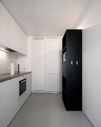 A narrow minimalist kitchen featuring seamless white slab cabinets, a matte black shelving column, and satin gray flooring.