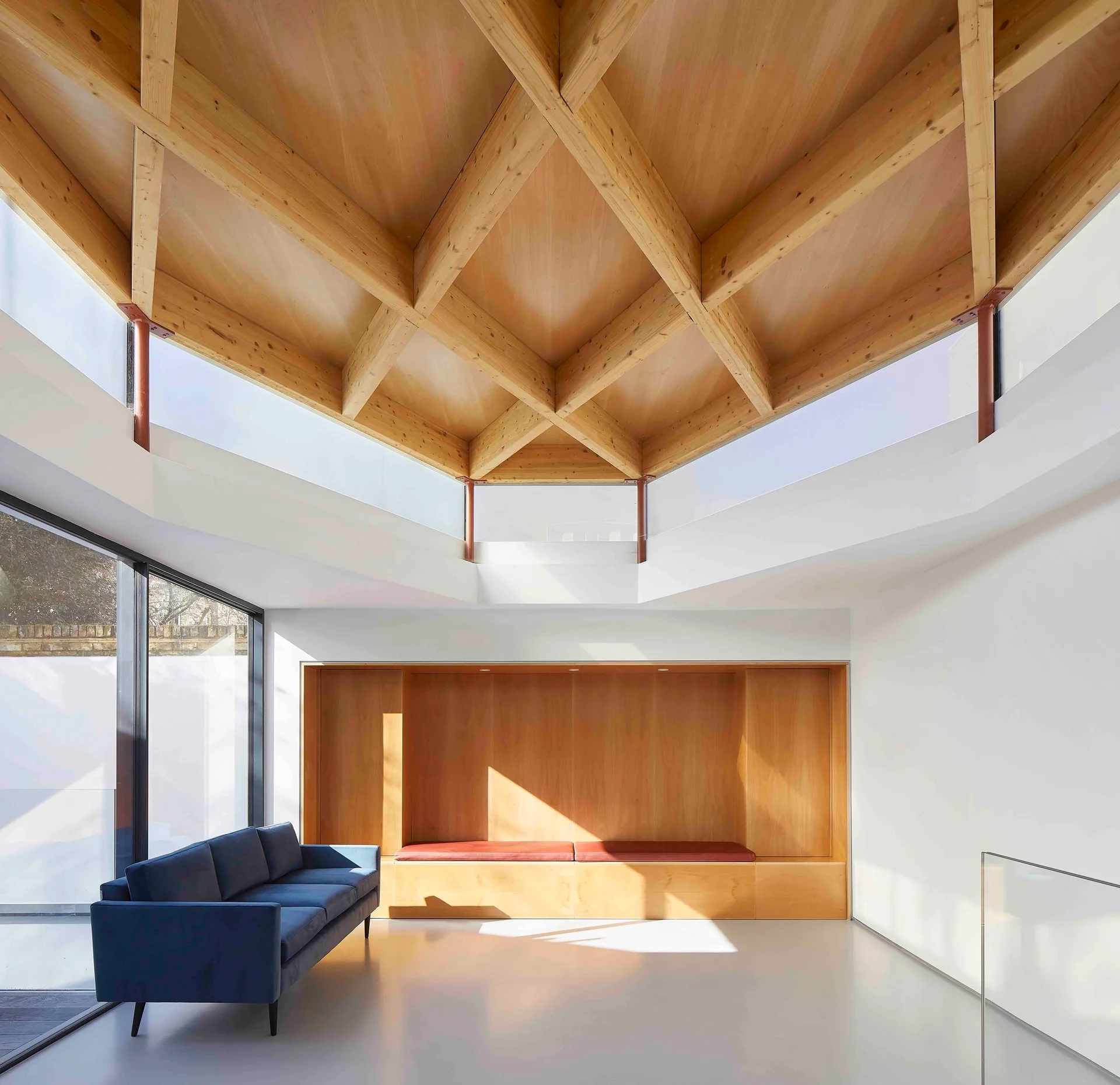 A minimalist living room features a geometric timber ceiling, a blue sofa, and a large wood built-in bench under clerestory windows.
