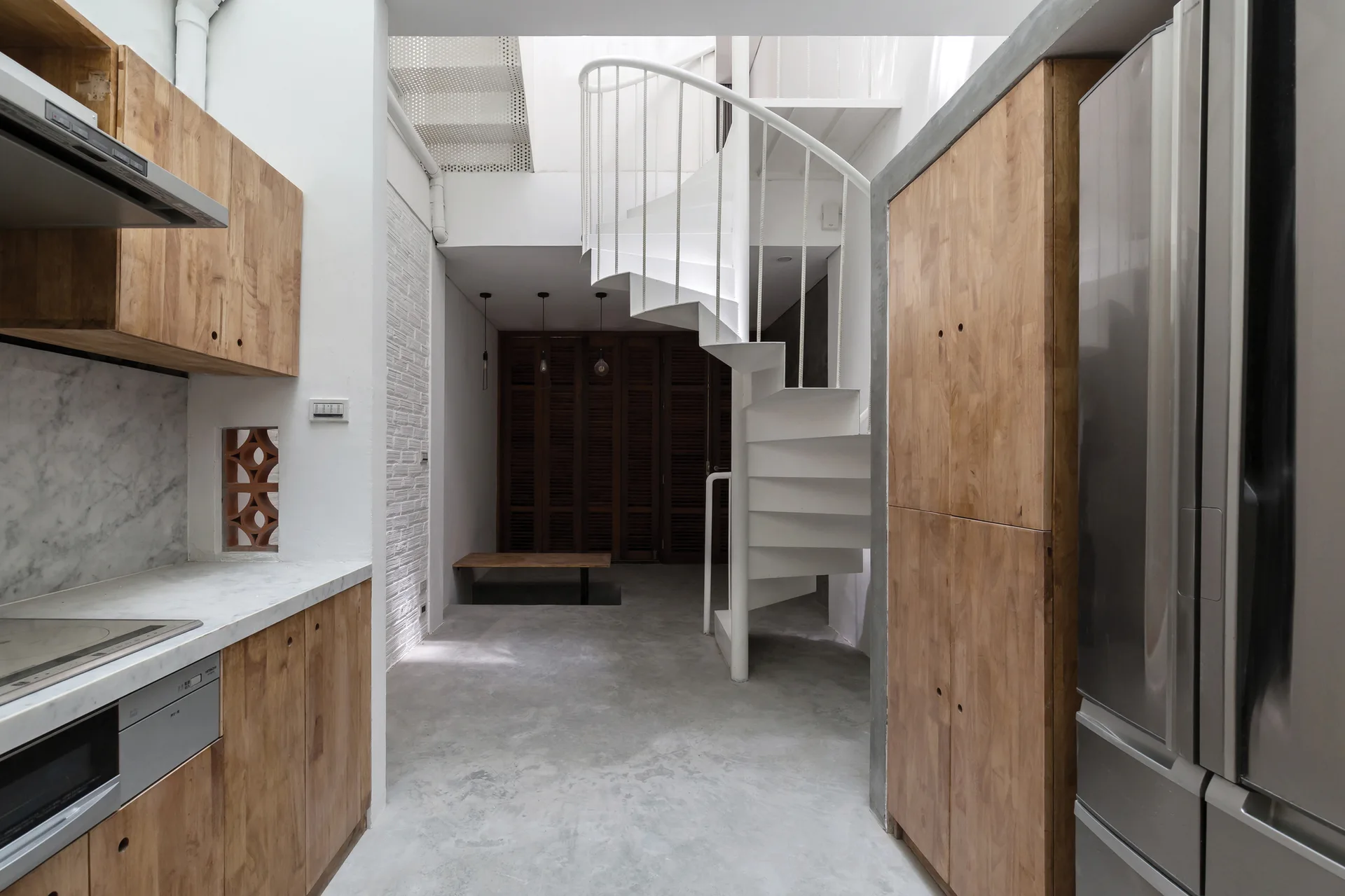 A modern minimalist kitchen featuring warm wooden cabinets, grey concrete flooring, and a white spiral staircase leading upward.